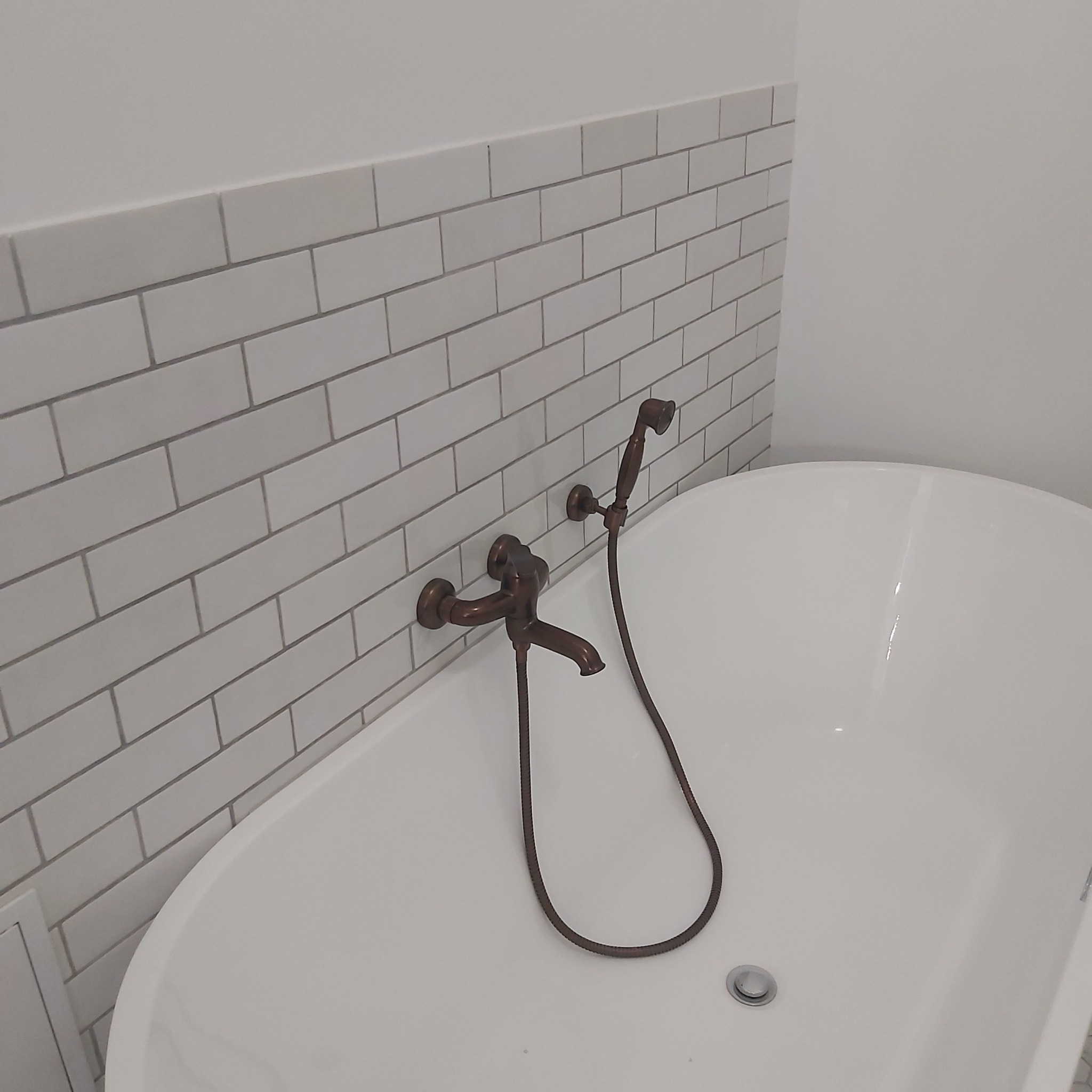 Biała wanna wolnostojąca z miedzianą armaturą i baterią prysznicową na tle ściany wyłożonej białymi, prostokątnymi płytkami imitującymi cegłę.