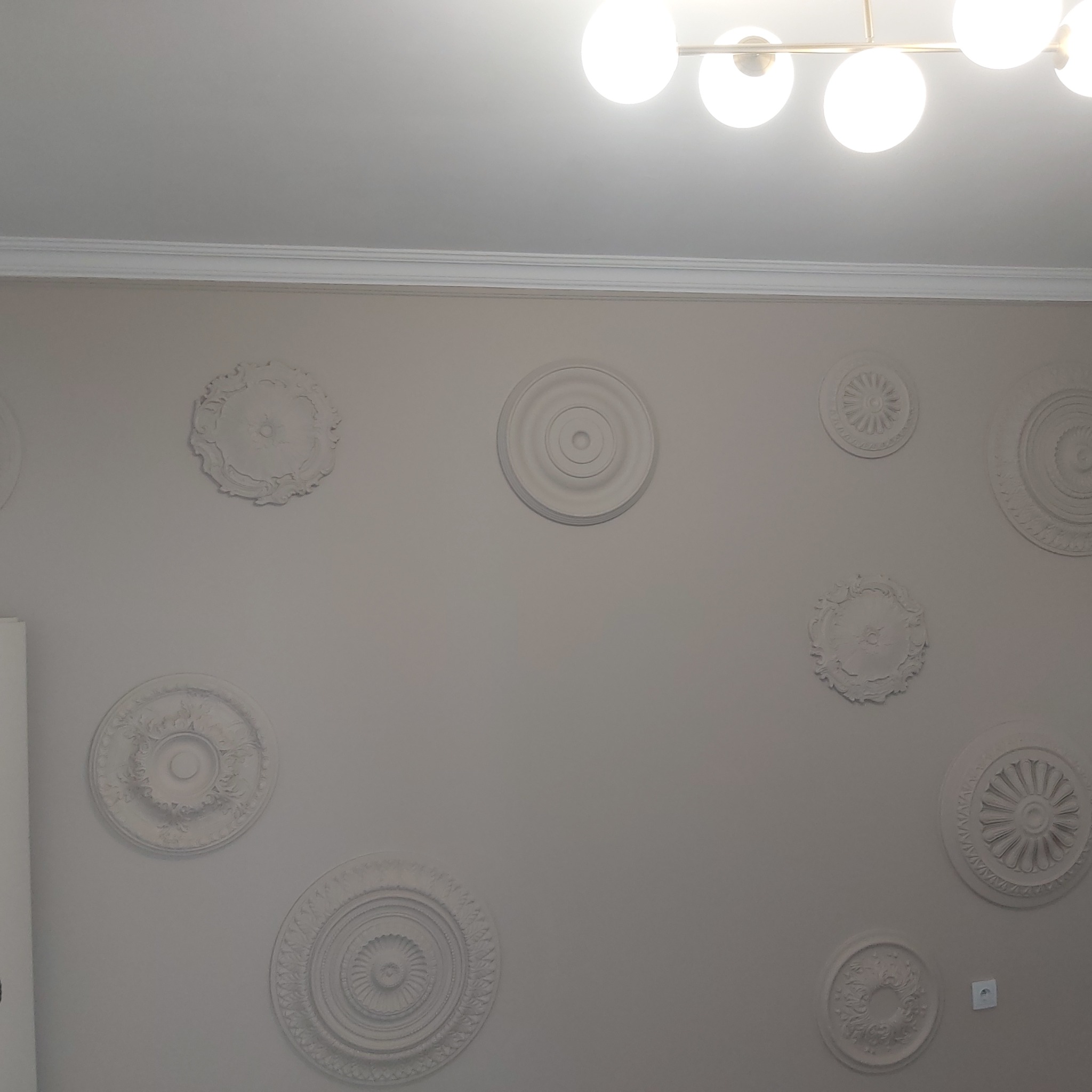 Szara ściana z dekoracyjnymi, okrągłymi sztukateriami o różnych wzorach, widoczne również listwy przysufitowe i nowoczesna lampa sufitowa z kulistymi kloszami.