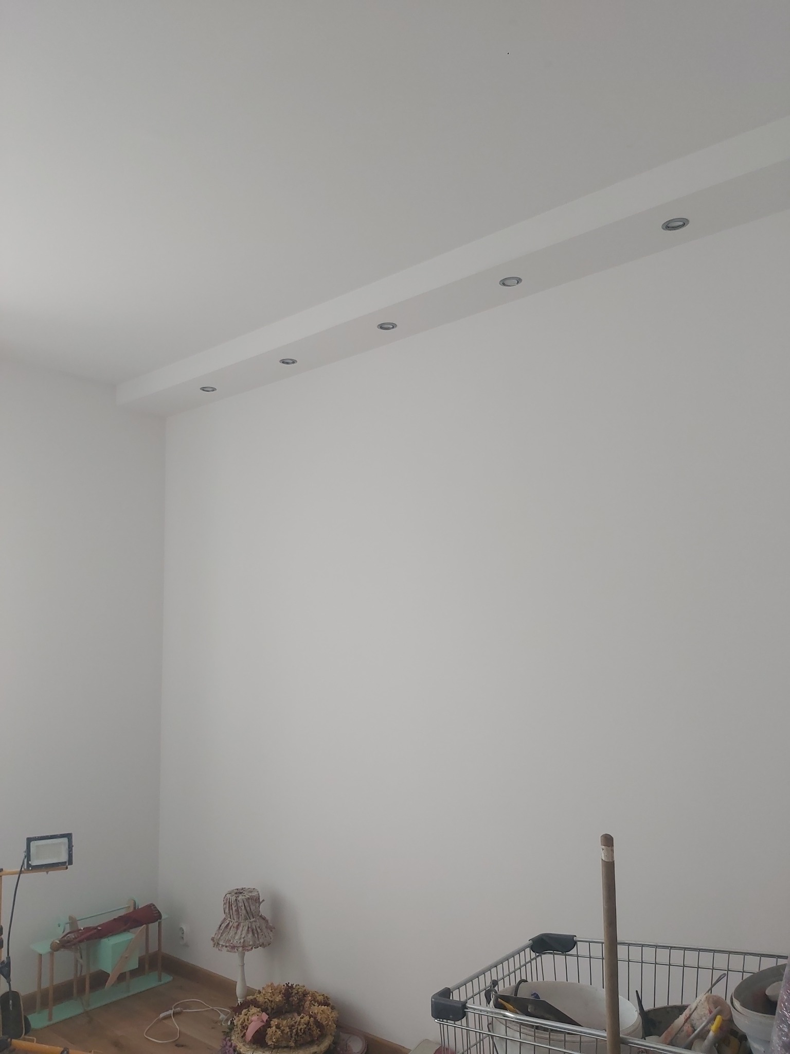 Wnętrze pokoju po malowaniu ścian na biało z zamontowanym oświetleniem sufitowym, widoczne narzędzia malarskie w wózku oraz dekoracje w rogu pomieszczenia.