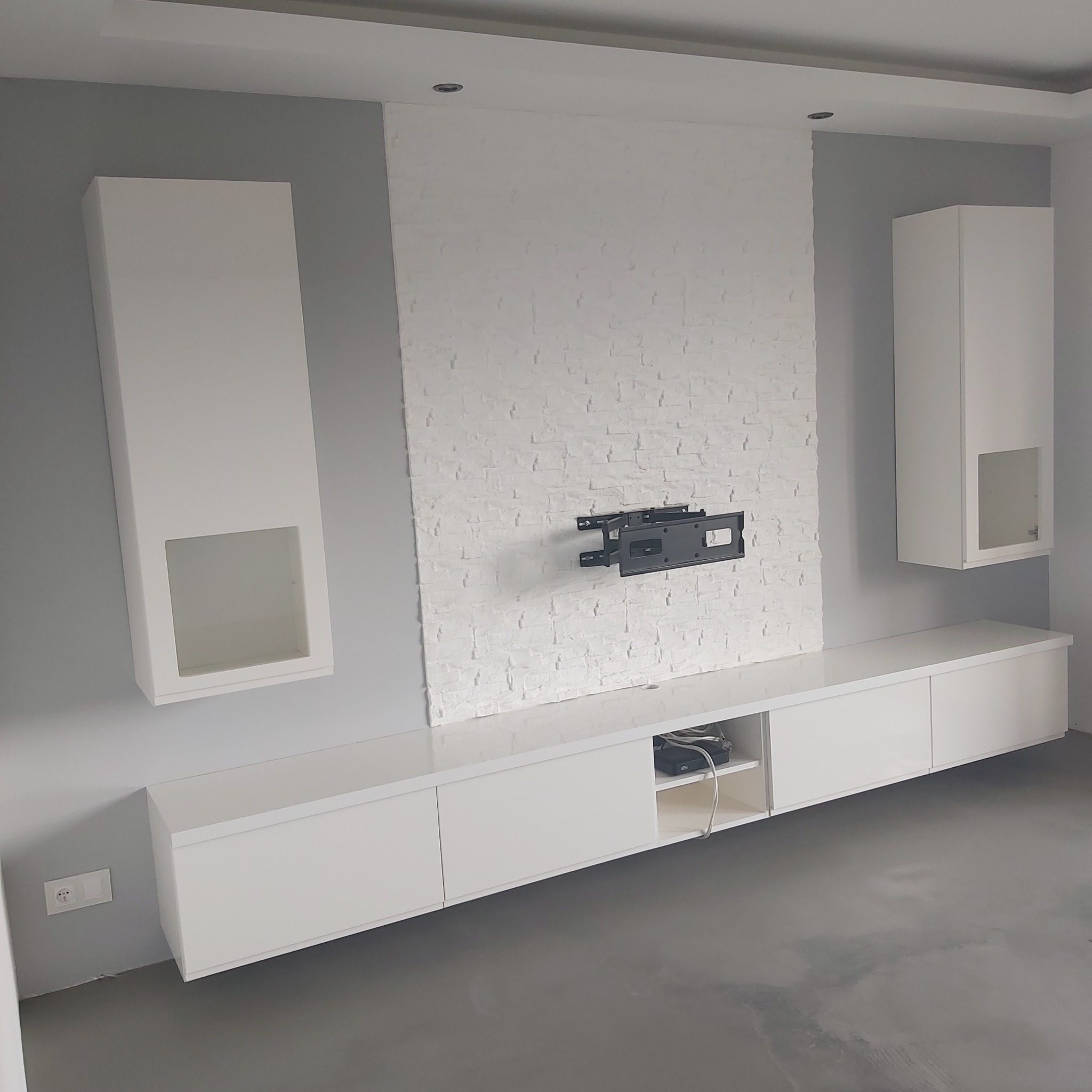 Nowoczesna aranżacja salonu z białymi meblami na wymiar, ściana akcentowa z dekoracyjnej białej cegły i uchwytem do telewizora, szara ściana boczna i szara podłoga.