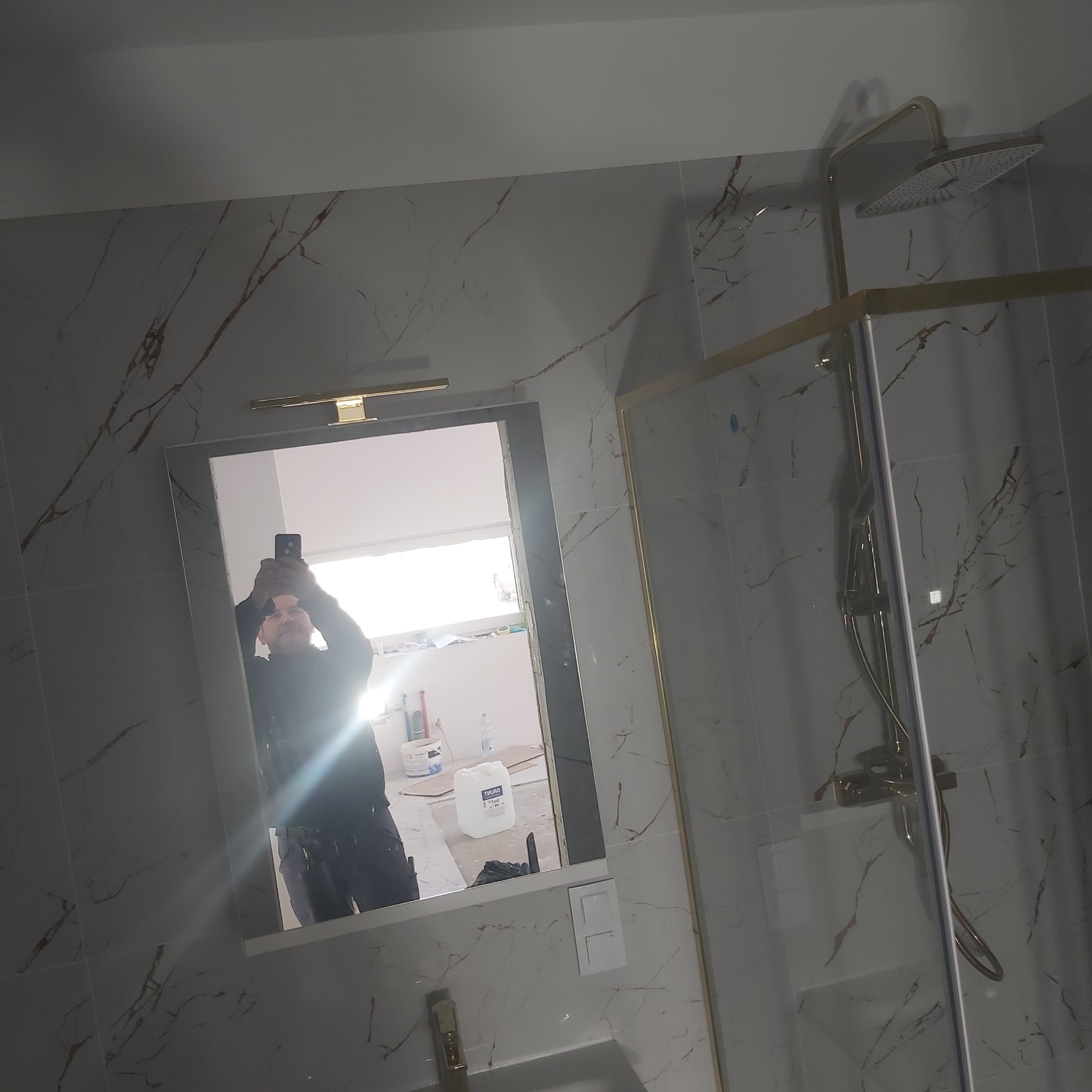 Wykończona łazienka z kabiną prysznicową typu walk-in, wykończona płytkami imitującymi marmur z żyłami w kolorze miedzi, widoczne odbicie osoby robiącej zdjęcie w lustrze.