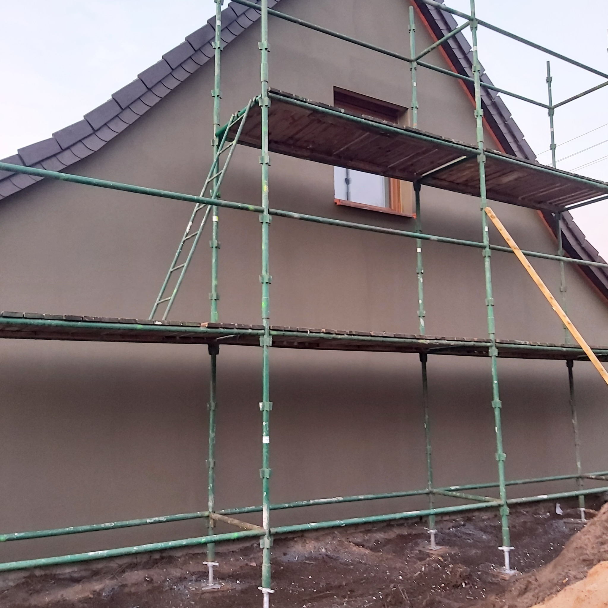 Dom jednorodzinny w trakcie prac elewacyjnych, widoczne rusztowanie, nałożona warstwa szarej gładzi, brązowe okno, dach pokryty ciemną dachówką.