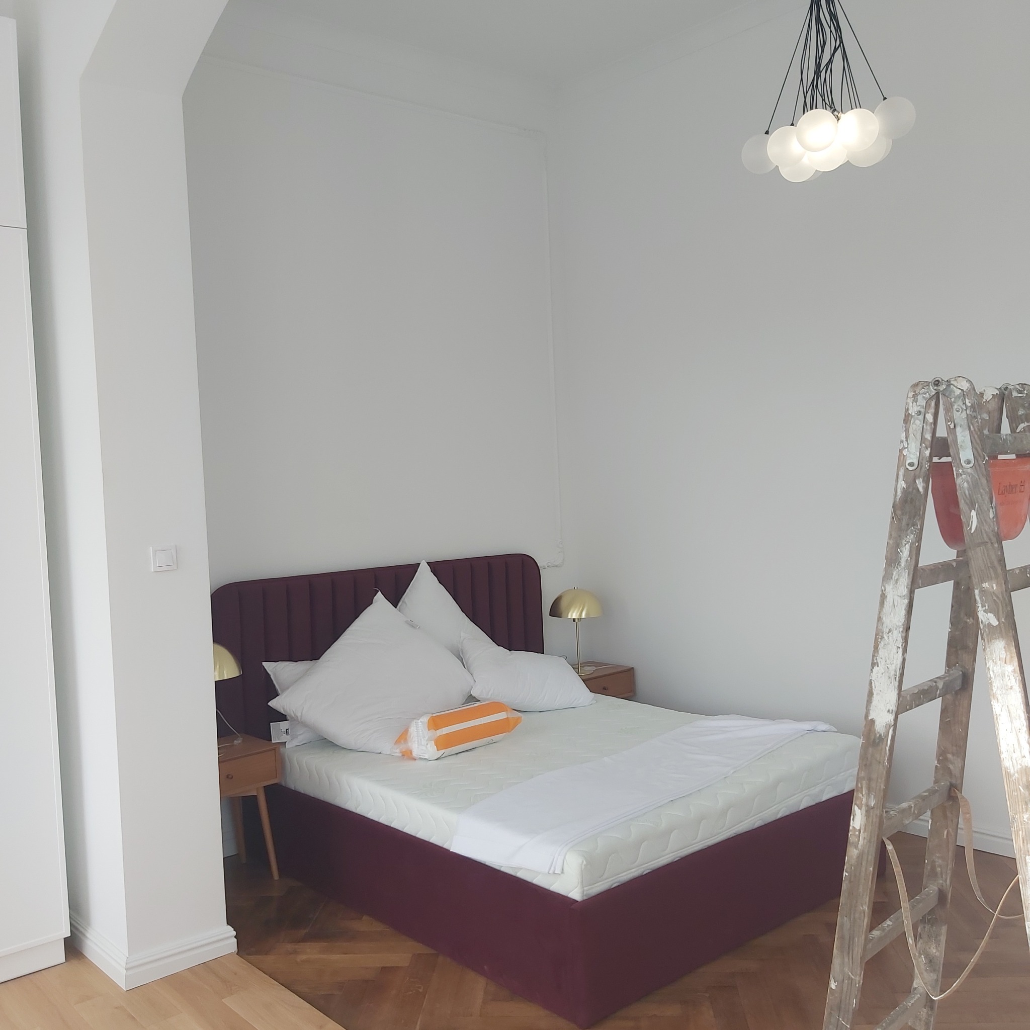 Pokój po odświeżeniu ścian, widoczna drabina malarska, burgundowe łóżko z białymi poduszkami i lampki nocne, parkiet na podłodze i nowoczesna lampa sufitowa.