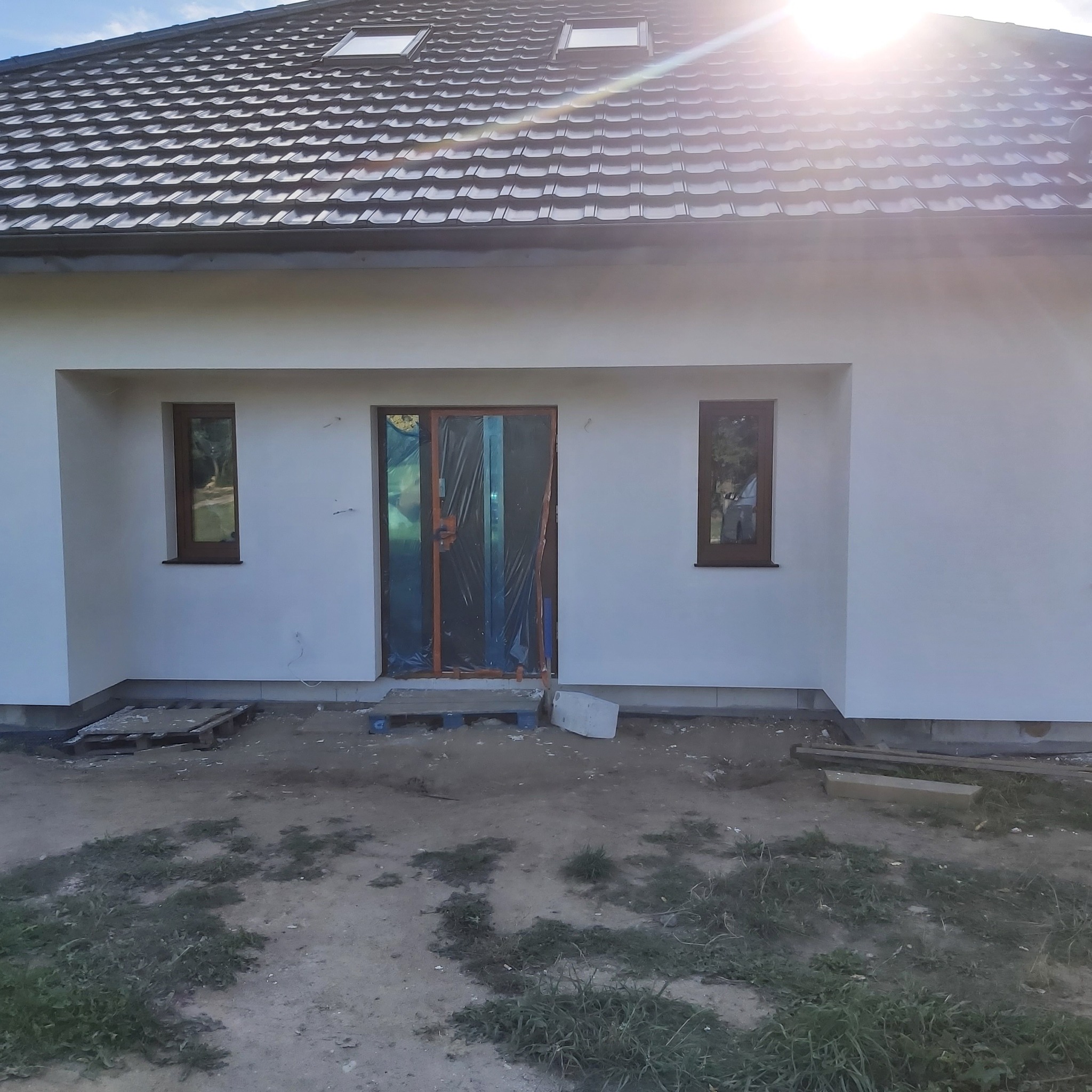 Fragment elewacji domu jednorodzinnego po nałożeniu gładzi, z oknami w brązowych ramach i zabezpieczonymi folią drzwiami wejściowymi, widoczne palety i resztki materiałów budowlanych na ziemi.