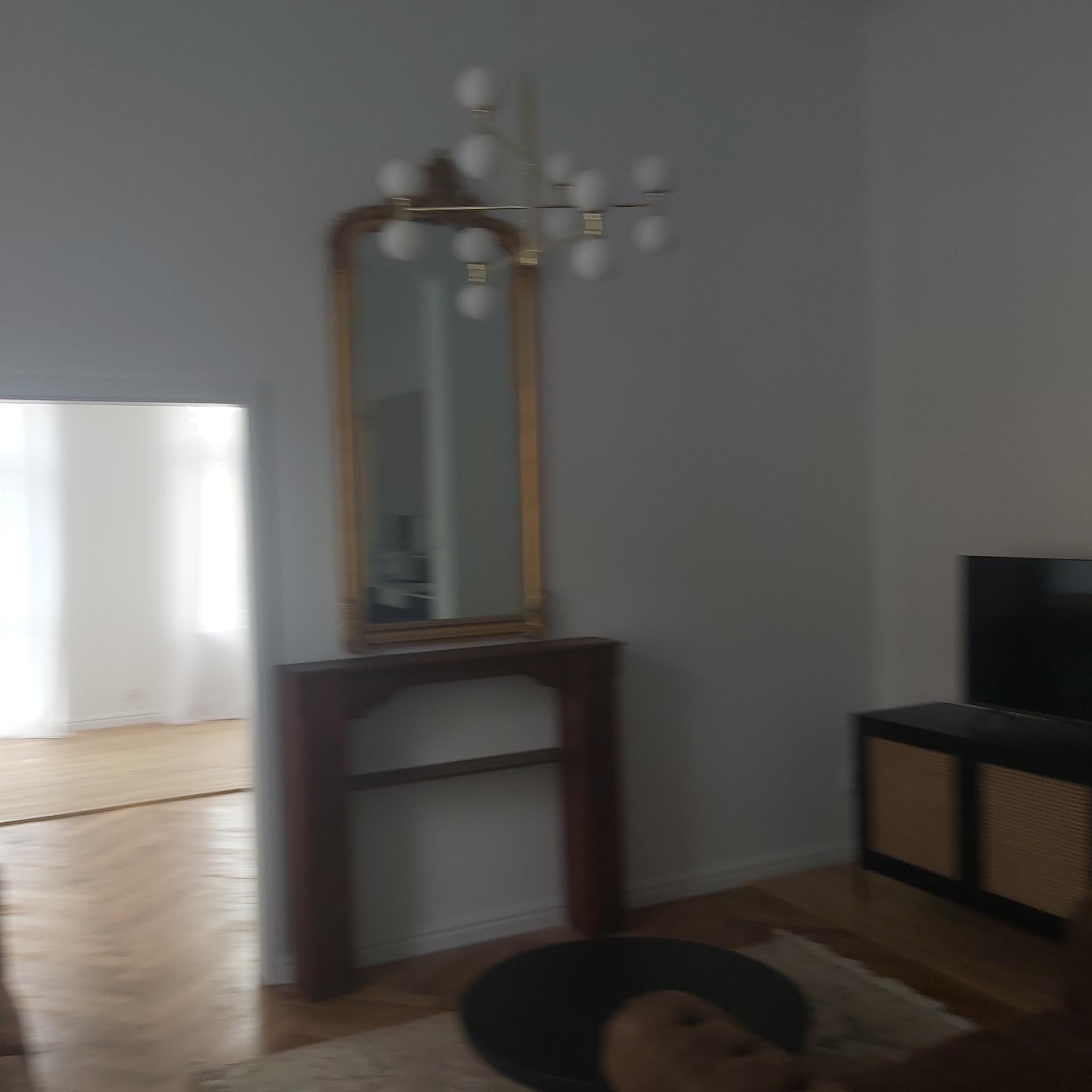 Jasne wnętrze mieszkania z drewnianą podłogą, lustrem w złotej ramie nad drewnianą konsolą i nowoczesną lampą sufitową.