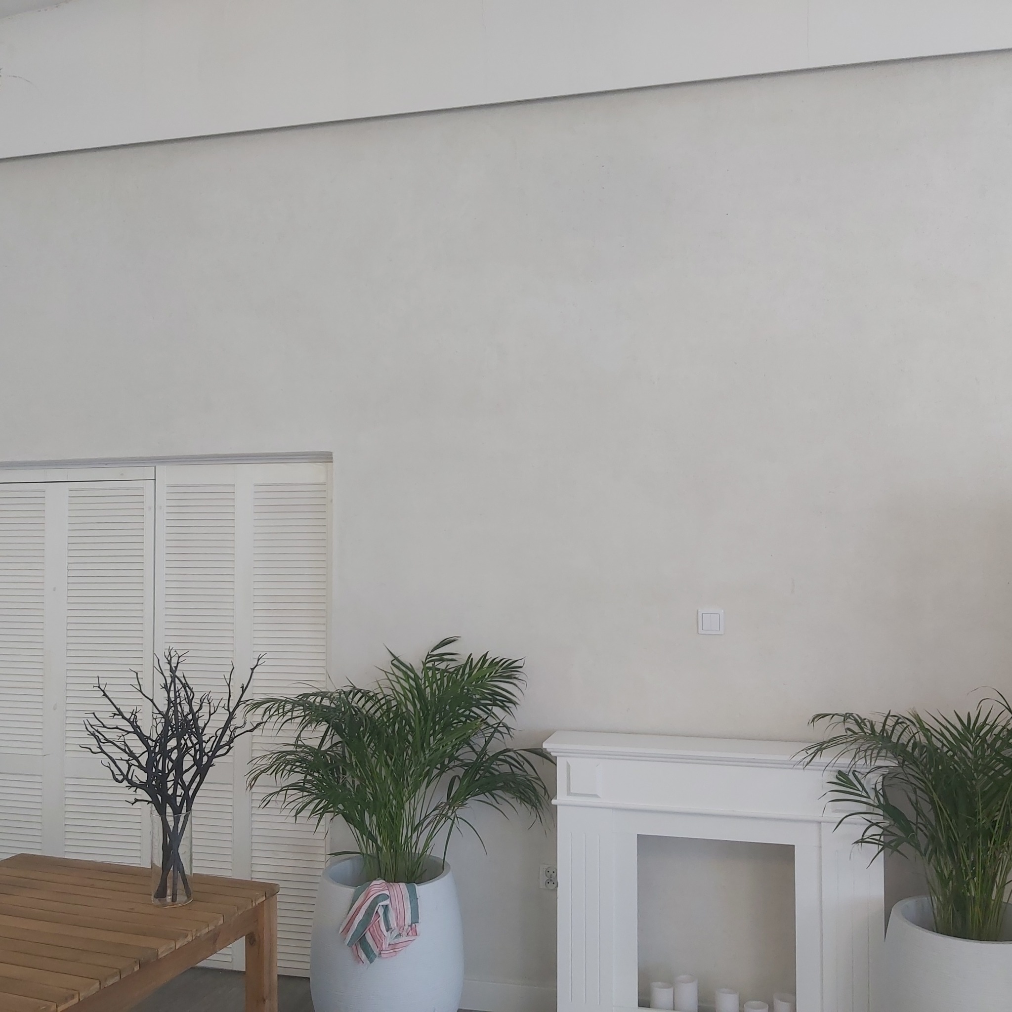 Jasne wnętrze z gładką ścianą, drewnianym stołem z czarnym bukietem w wazonie, doniczkowymi palmami i białą atrapą kominka z świecami.