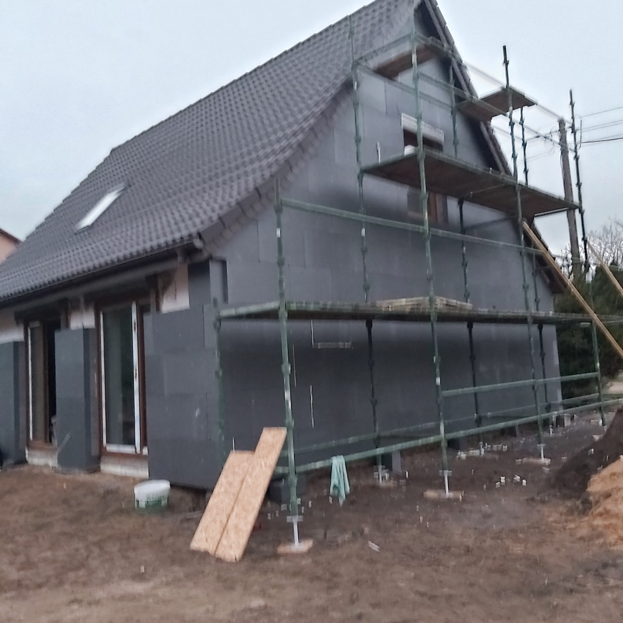 Dom jednorodzinny w trakcie ocieplania elewacji, widoczne szare płyty izolacyjne, rusztowanie, okna z brązowymi ramami oraz fragment dachu z dachówką.