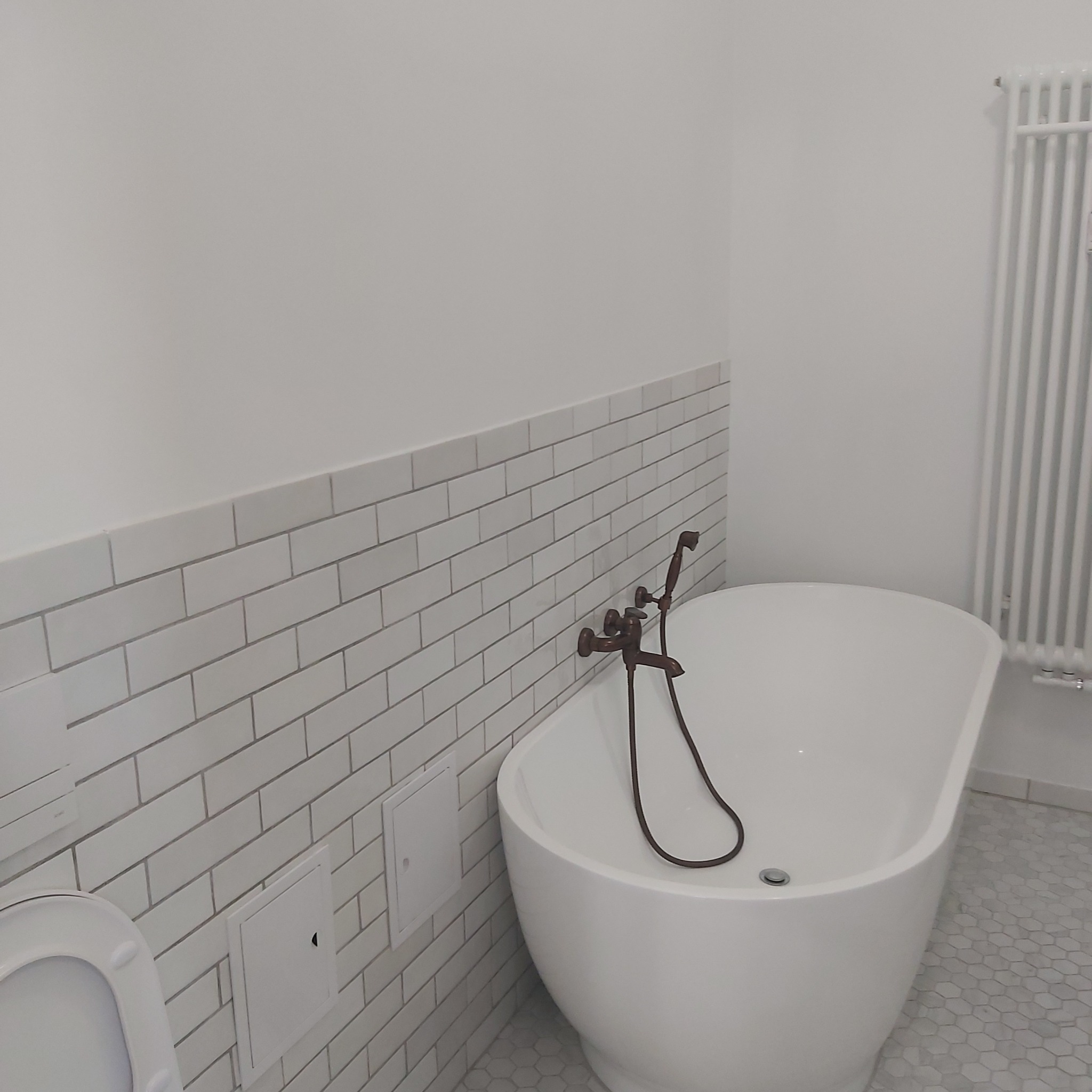 Biała wanna wolnostojąca z brązową armaturą na tle ściany wyłożonej białymi, prostokątnymi płytkami imitującymi cegłę, z widocznym fragmentem toalety i grzejnika.