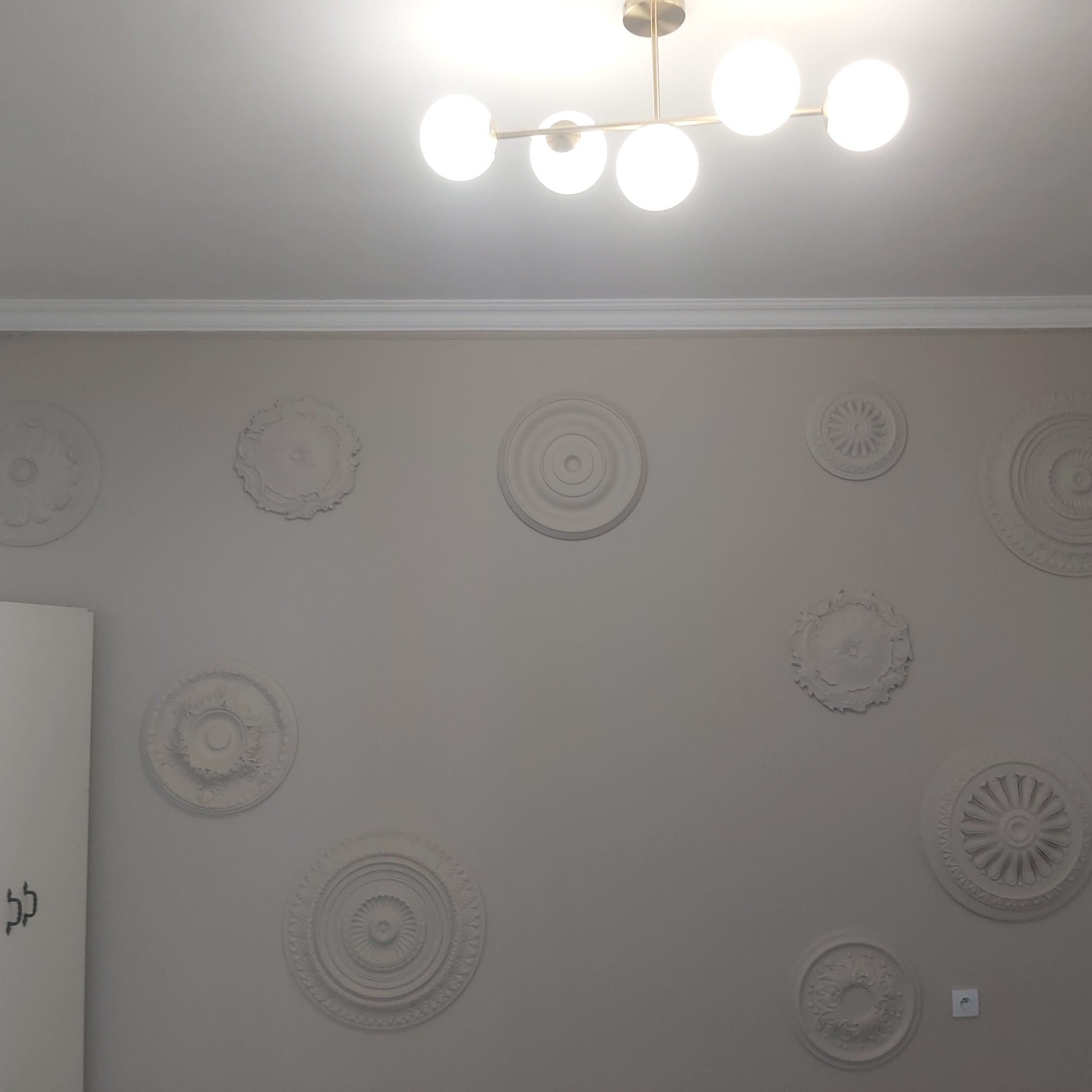 Ściana w pokoju z dekoracyjnymi, okrągłymi rozetami gipsowymi o różnych wzorach, oświetlona nowoczesną lampą sufitową z kulistymi kloszami.