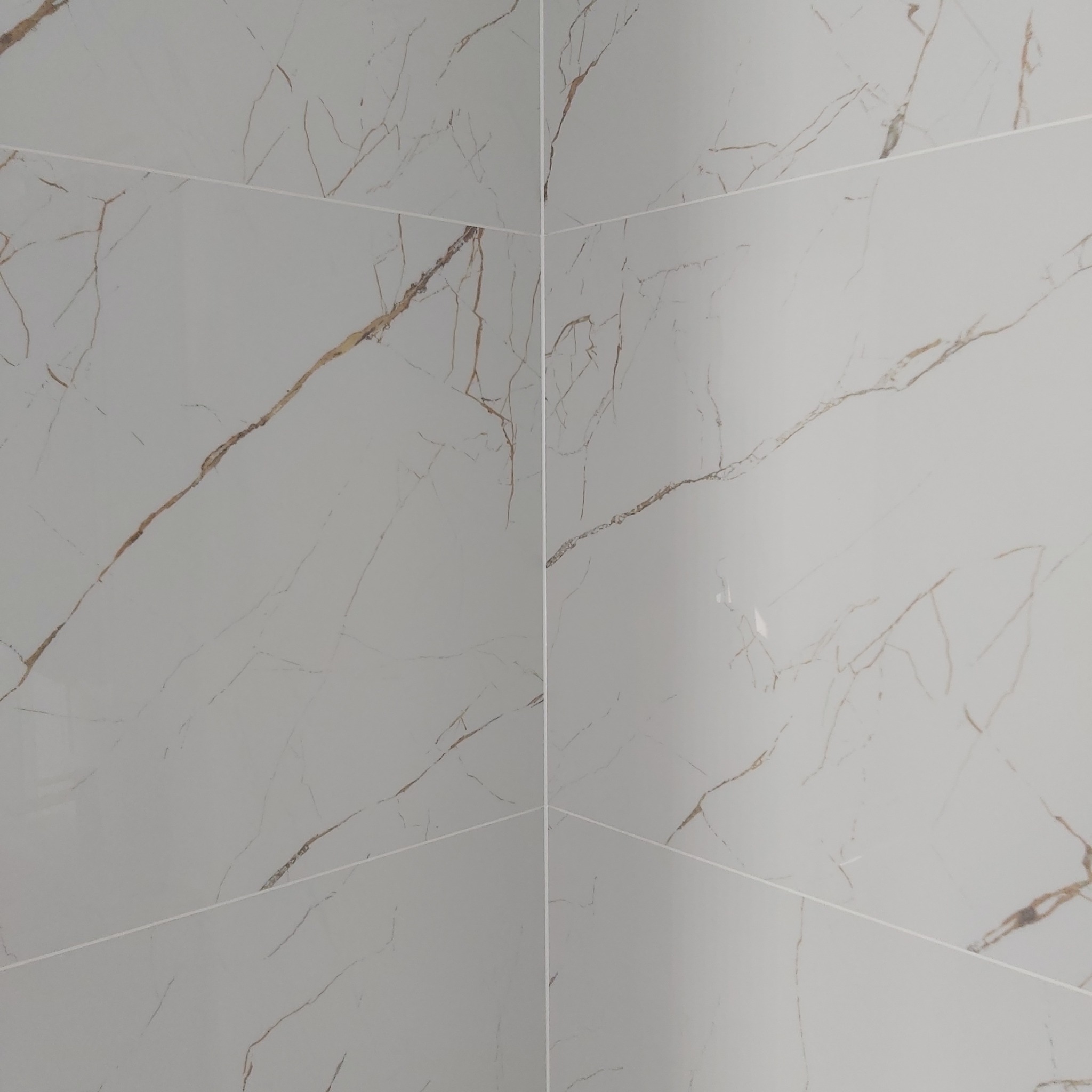 Ściana wyłożona jasnoszarymi płytkami imitującymi marmur z wyraźnymi złotymi żyłami, widoczne fugi między płytkami.