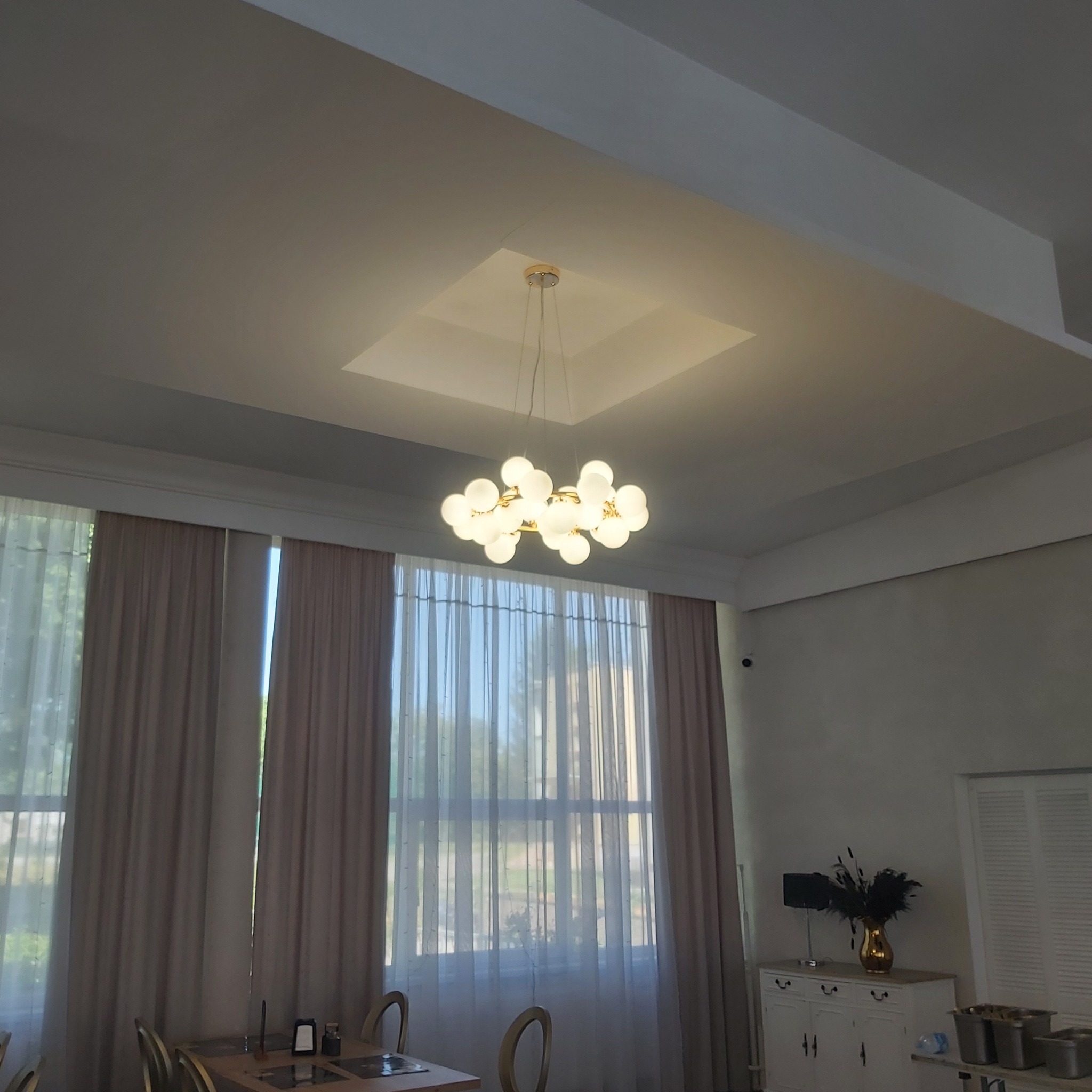 Nowoczesny salon z kremowymi zasłonami, białą komodą z dekoracjami i designerską lampą sufitową z kulistymi kloszami.