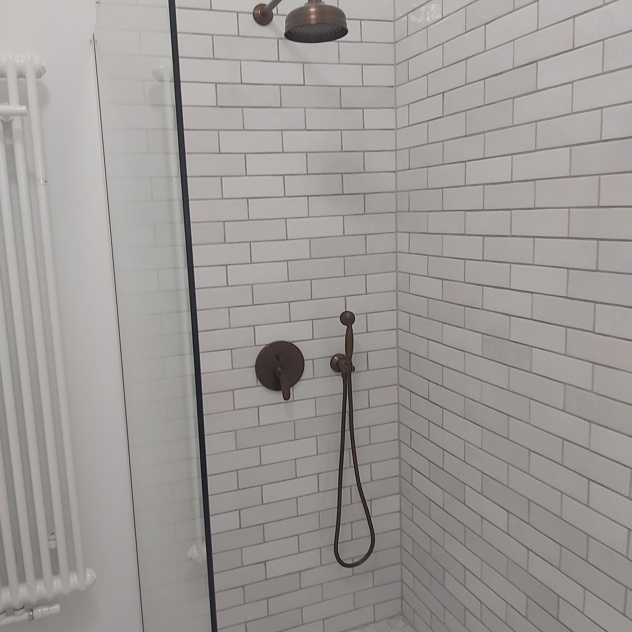 Wykończona kabina prysznicowa z białymi, podłużnymi płytkami ułożonymi cegiełkowo, brązowa armatura, szklane drzwi kabiny i biały grzejnik drabinkowy z boku.