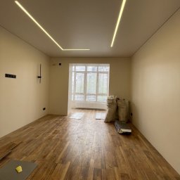 SWIWT SPÓŁKA Z OGRANICZONĄ ODPOWIEDZIALNOŚCIĄ - Pokój w trakcie wykańczania: panele podłogowe ułożone, na suficie geometryczne oświetlenie LED, worki z materiałami budowlanymi w rogu pomieszczenia, widok na okno.
