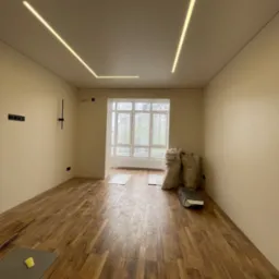 Pokój w trakcie wykańczania: panele podłogowe ułożone, na suficie geometryczne oświetlenie LED, worki z materiałami budowlanymi w rogu pomieszczenia, widok na okno.