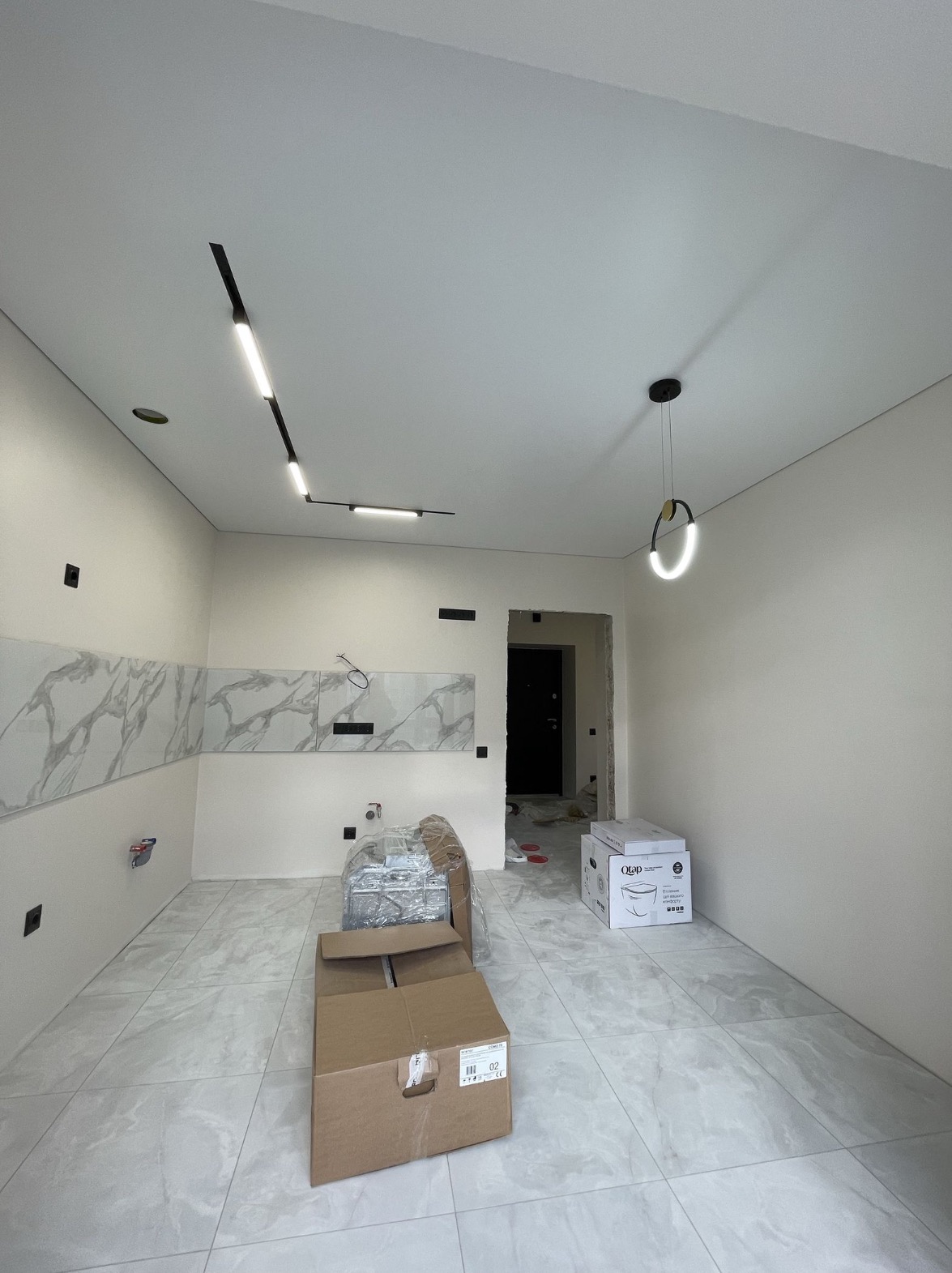 Wnętrze w trakcie prac wykończeniowych: ułożone płytki imitujące marmur na ścianie i podłodze, widoczne punkty elektryczne, kartony z wyposażeniem oraz nowoczesne oświetlenie - listwa LED i okrągła...