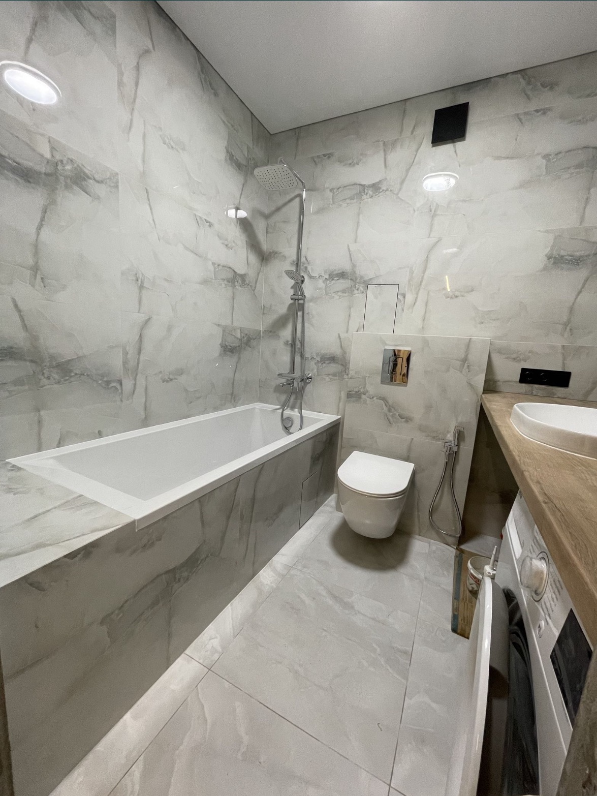 Nowoczesna łazienka w odcieniach szarości z marmurowymi płytkami, białą wanną, wiszącą toaletą, umywalką z drewnianym blatem i pralką, oświetlona punktowymi lampami sufitowymi.