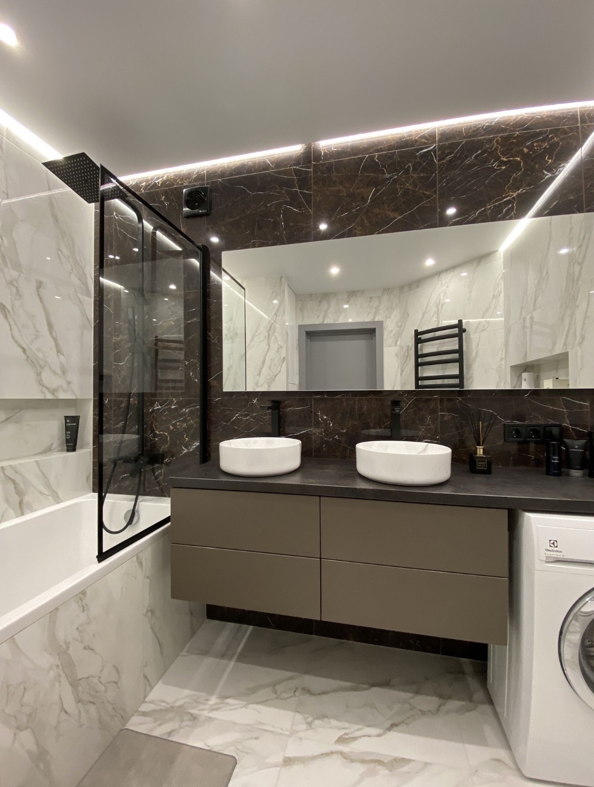 Nowoczesna łazienka z dwoma białymi umywalkami nablatowymi, czarną armaturą, kabiną prysznicową typu walk-in z czarnymi profilami, marmurowymi płytkami w odcieniach bieli i brązu...