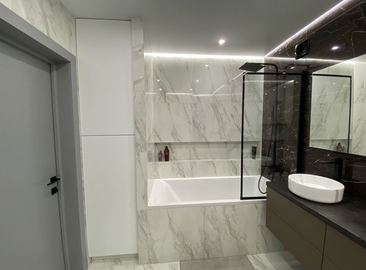 Nowoczesna łazienka z wanną obudowaną marmurowymi płytkami, czarną kabiną prysznicową typu walk-in i umywalką nablatową na brązowej szafce, oświetlenie LED podkreśla marmurową teksturę.