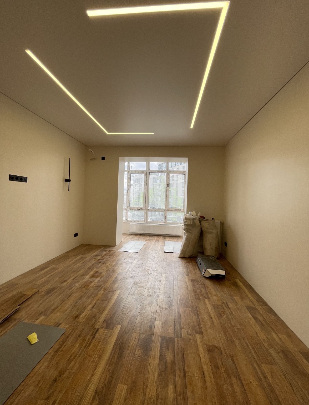 Pokój w trakcie wykańczania: panele podłogowe ułożone, na suficie geometryczne oświetlenie LED, worki z materiałami budowlanymi w rogu pomieszczenia, widok na okno.