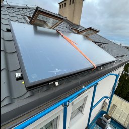SANITARINSTAL Irena Rygielska - Montaż panelu słonecznego na dachu z dachówką ceramiczną, widok z podnośnika, pomarańczowy pas zabezpieczający, otwarte okna dachowe, pochmurne niebo.