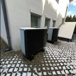 Dwie czarne pompy ciepła Daikin Altherma 3 R ECH2O, stojące na kostce brukowej pokrytej śniegiem przed jasnym budynkiem z oknami i rynną spustową.