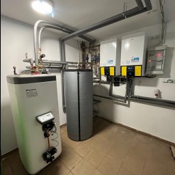 SANITARINSTAL Irena Rygielska - Wnętrze kotłowni z dwoma białymi jednostkami grzewczymi Daikin zamontowanymi na ścianie, obok pionowy zbiornik akumulacyjny w szarej izolacji i drugi, większy zbiornik Daikin z widocznym panelem...