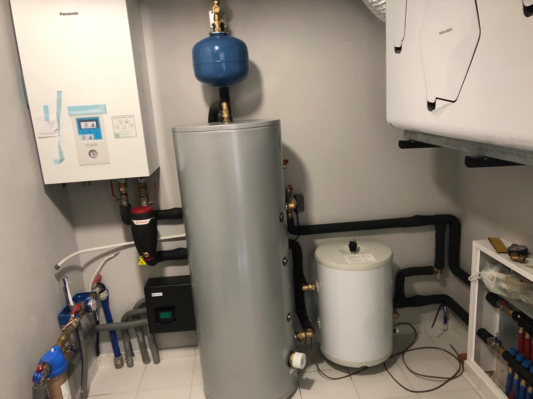 Kotłownia z piecem gazowym Panasonic, zbiornikiem akumulacyjnym oraz osprzętem hydraulicznym, widoczne rury i zawory.