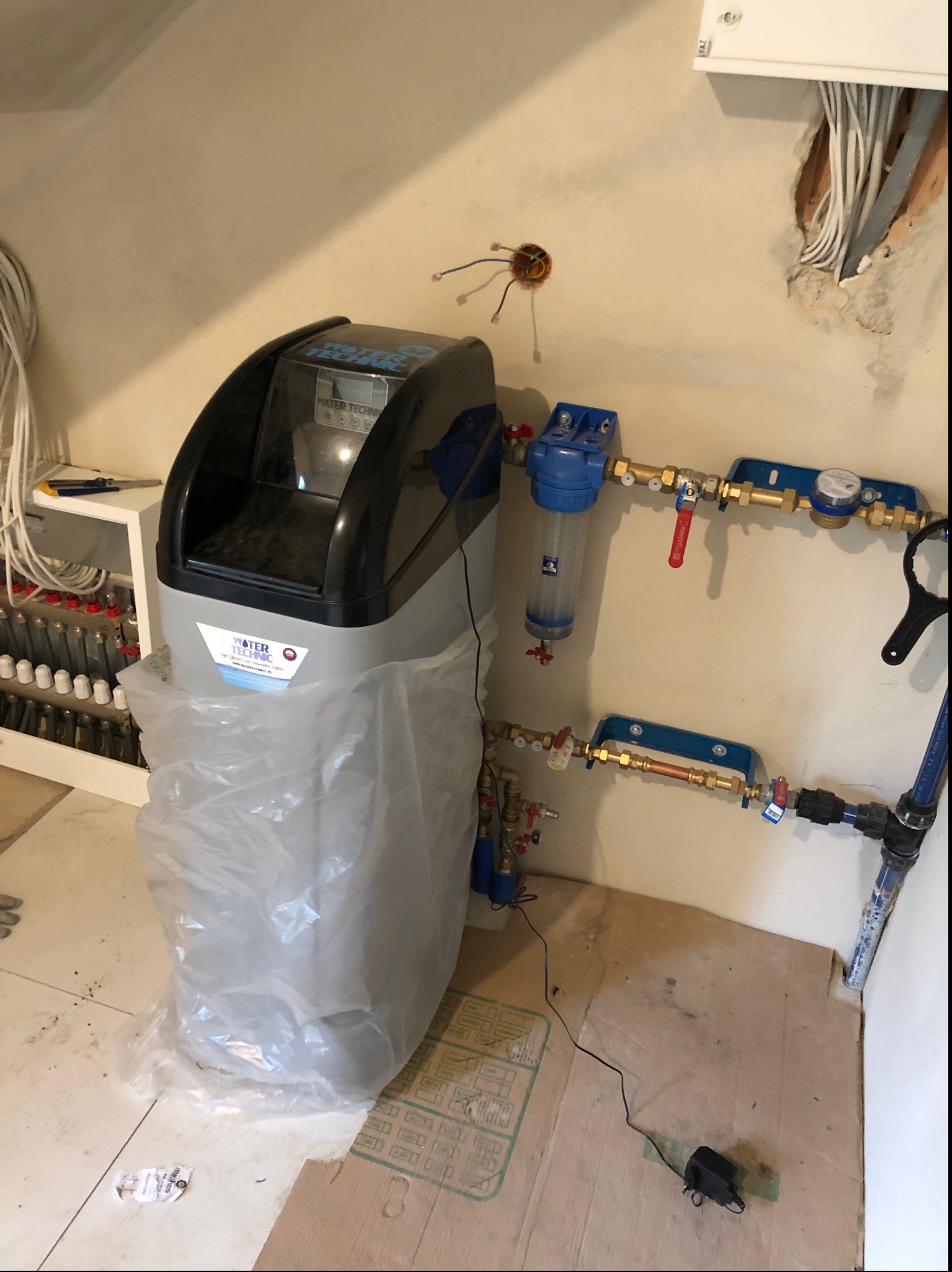 Szara stacja uzdatniania wody marki Water Technic, częściowo owinięta w folię ochronną, podłączona do instalacji wodnej z filtrami i licznikami, na tle surowej ściany z wystającymi kablami.