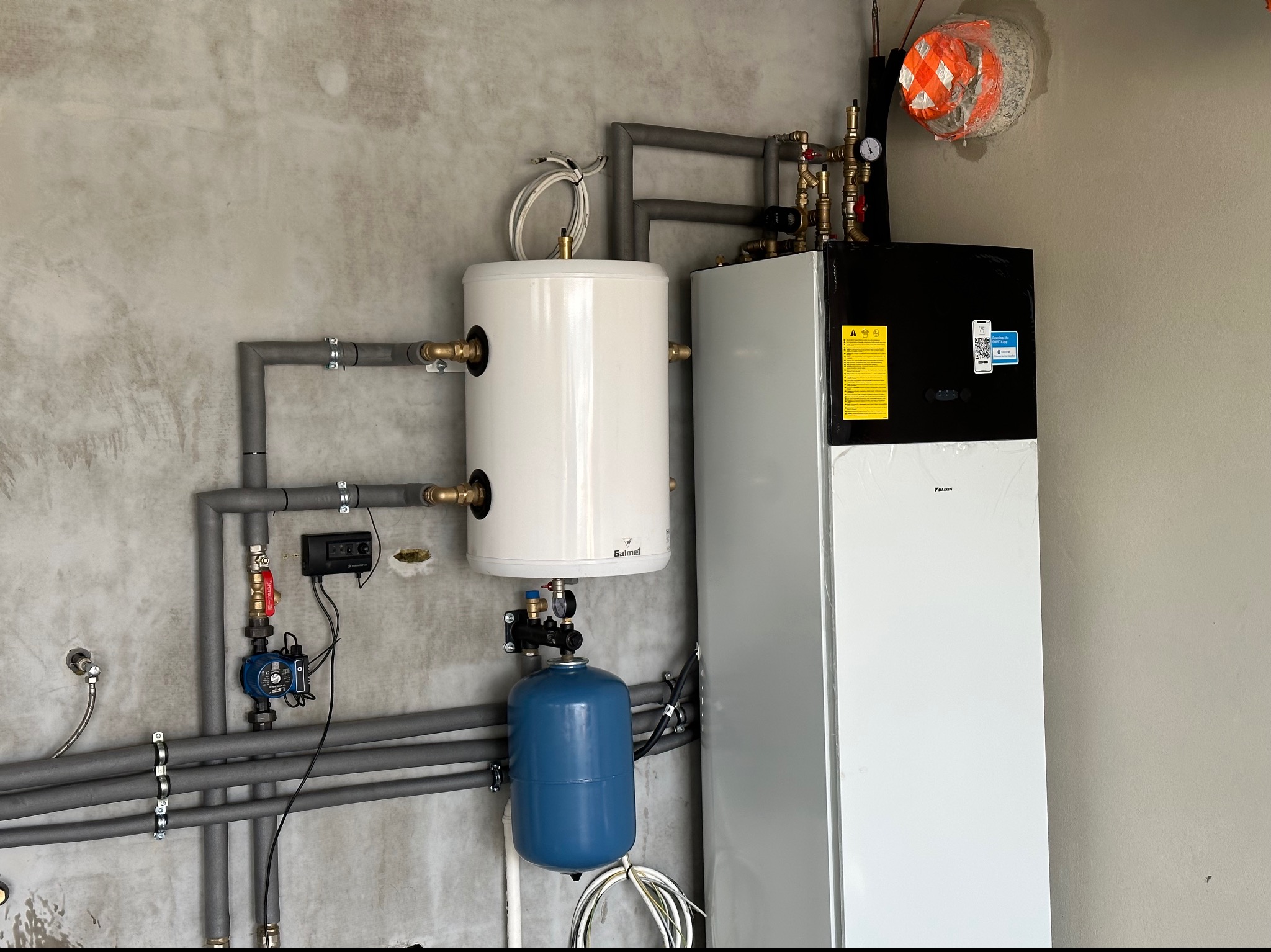 Zestaw urządzeń grzewczych: biały zbiornik Galmet połączony z szarymi rurami, niebieskie naczynie wzbiorcze i jednostka Daikin w folii ochronnej na tle surowej ściany.