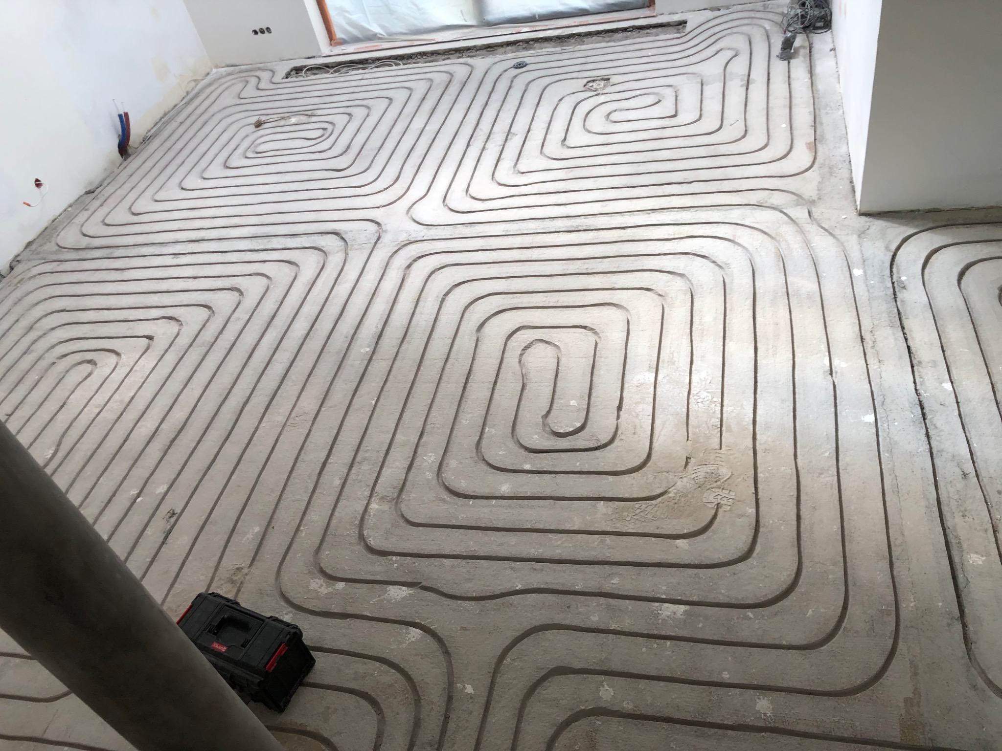 Układanie rur ogrzewania podłogowego w jastrychu, widoczna czarna skrzynka narzędziowa i fragment rury kanalizacyjnej.