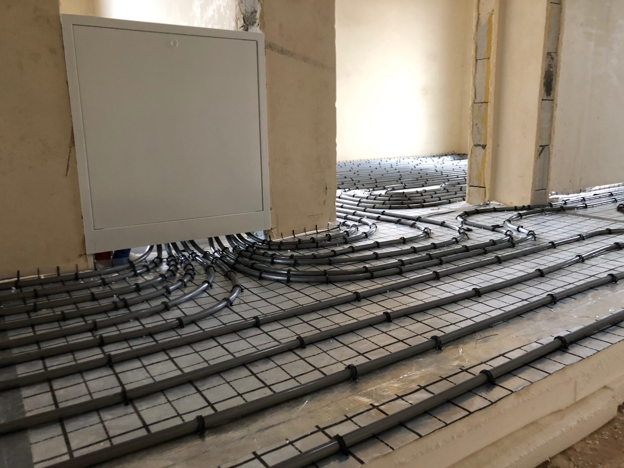 Szare rury ogrzewania podłogowego, mocowane do siatki na izolacji, prowadzone do białej skrzynki rozdzielczej w surowym wnętrzu budynku.
