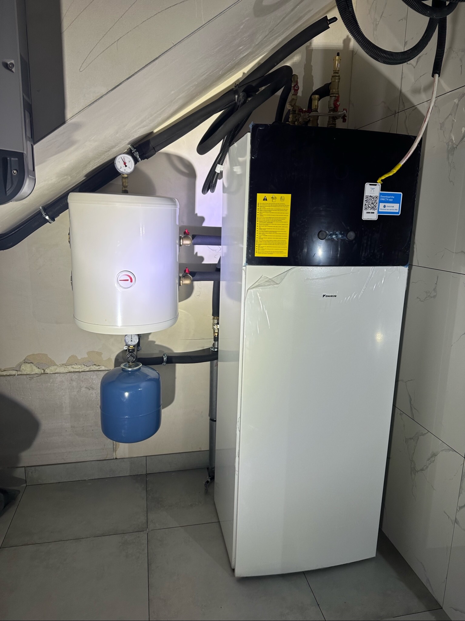 Biała pompa ciepła Daikin z podłączonym zasobnikiem CWU, widoczne rury instalacyjne, manometr i naklejki informacyjne, całość zamontowana w pomieszczeniu z szarymi płytkami.