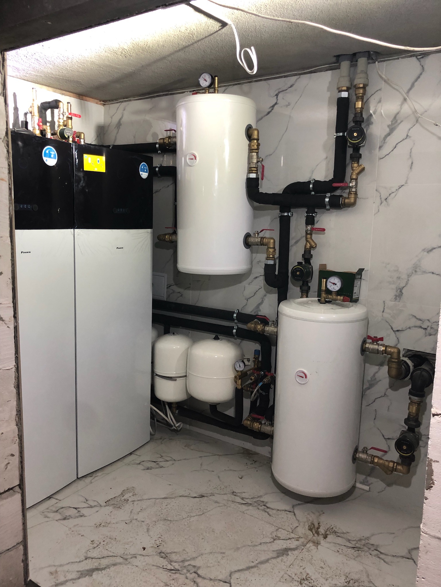Zainstalowany system grzewczy Daikin z dwoma zbiornikami akumulacyjnymi i naczyniami przeponowymi w pomieszczeniu z marmurowymi płytkami na ścianach i podłodze.