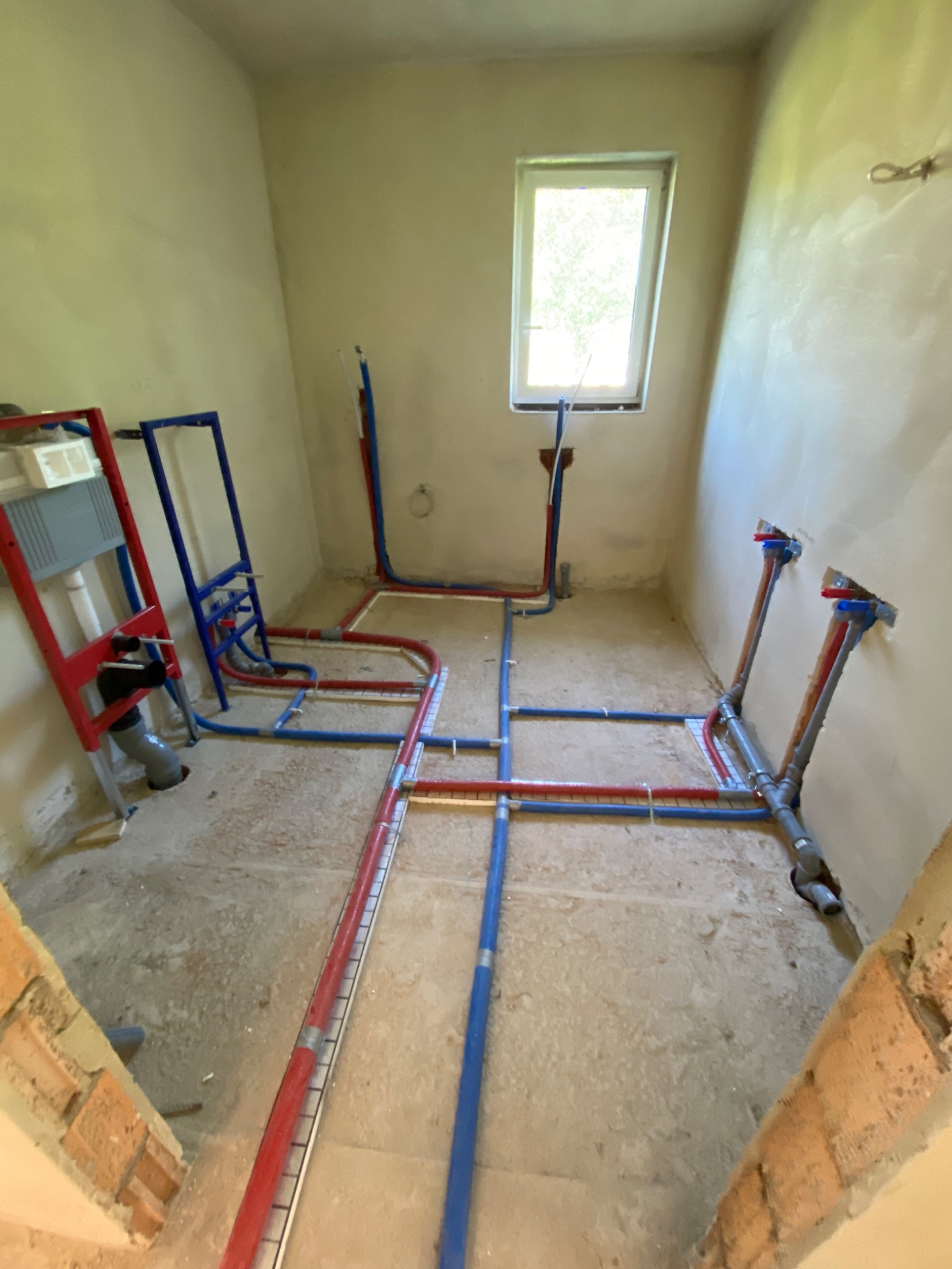 Surowa łazienka w trakcie instalacji wodnej i kanalizacyjnej; widoczne rury w kolorze czerwonym i niebieskim, stelaże podtynkowe WC oraz wyprowadzenia wody na ścianach.