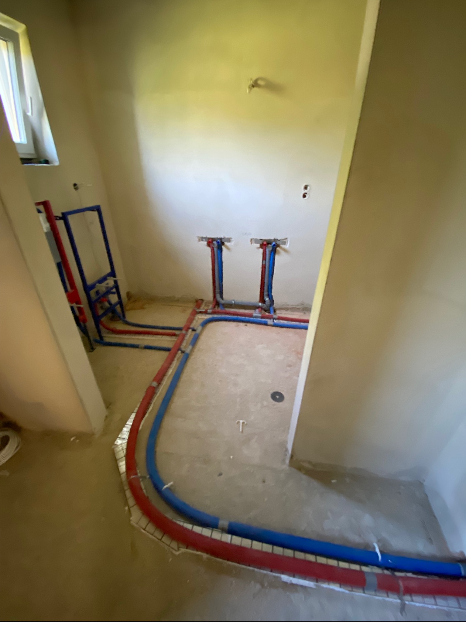 Surowe wnętrze łazienki w trakcie prac hydraulicznych: widoczne rury grzewcze ułożone w podłodze, pionowe przyłącza do armatury i stelaż podtynkowy WC.