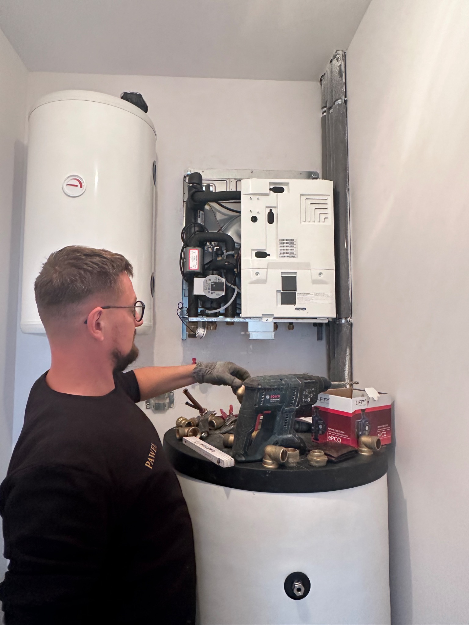 Instalator w okularach, w trakcie montażu pieca gazowego, widoczne narzędzia, złączki hydrauliczne i biały zasobnik ciepłej wody.