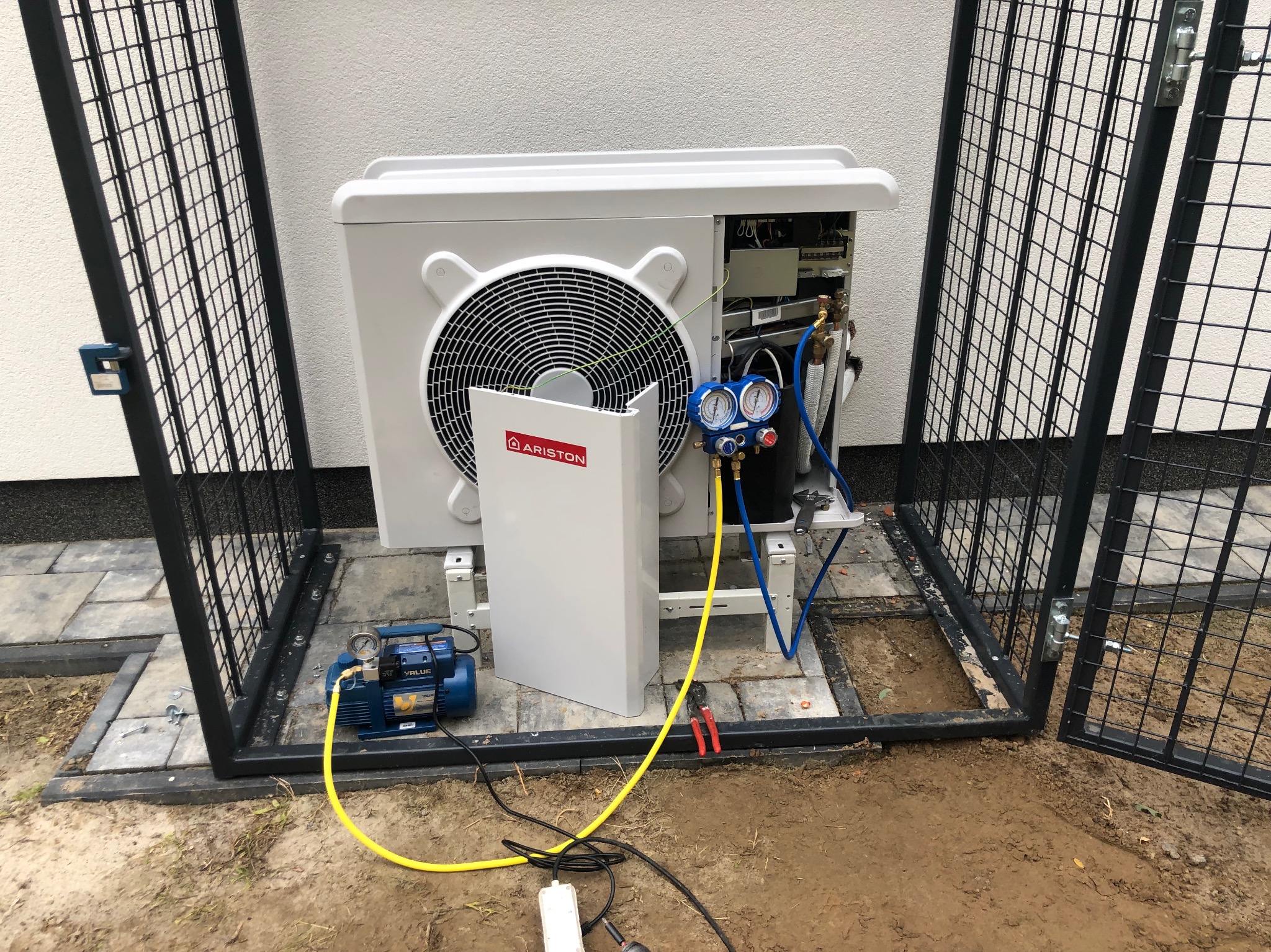 Biała pompa ciepła marki Ariston, zabezpieczona metalową klatką, podłączona do niebieskiej pompy próżniowej, manometry i przewody w trakcie instalacji na zewnątrz budynku.