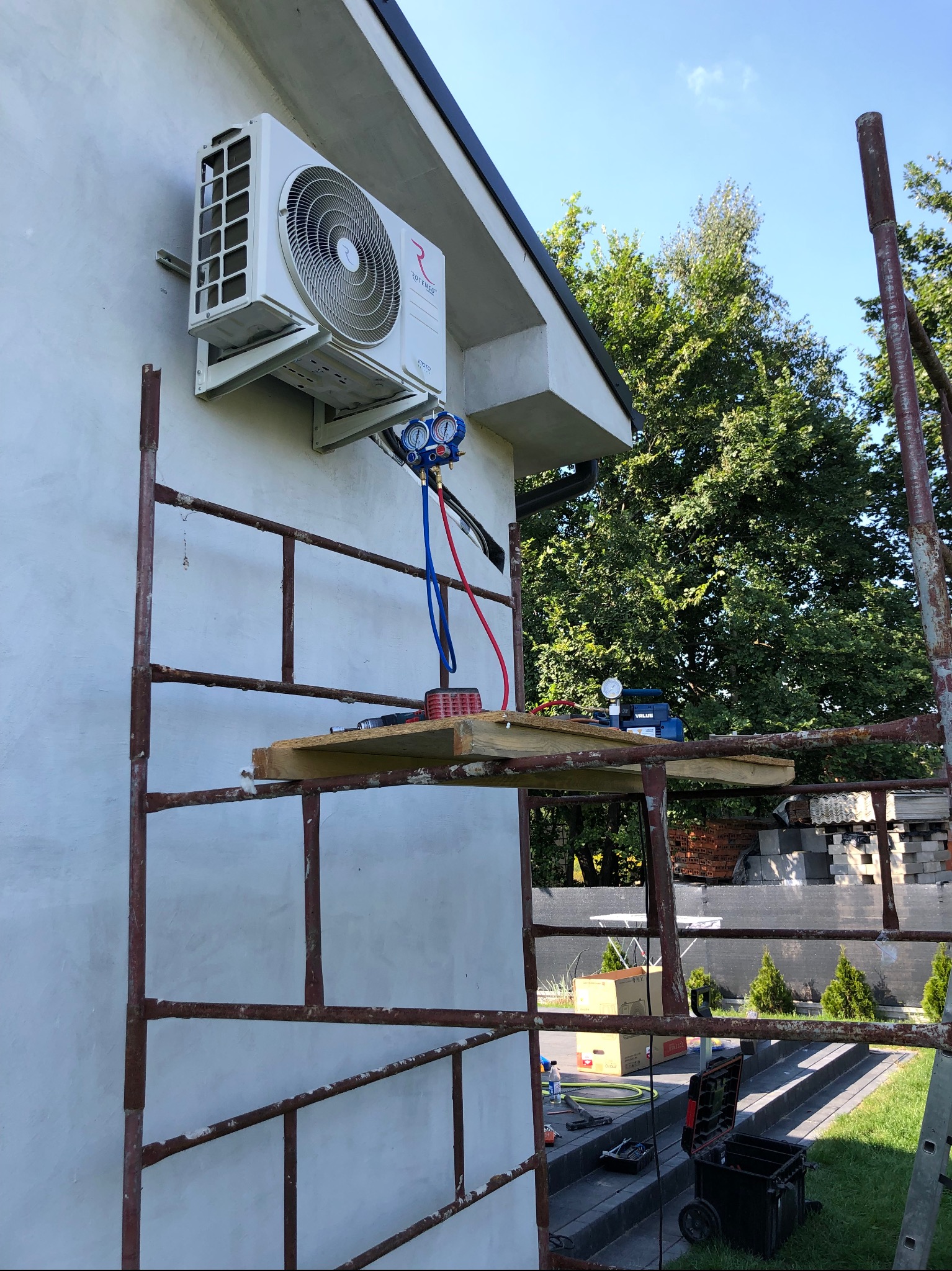 Biała jednostka zewnętrzna klimatyzacji zamontowana na elewacji budynku, widoczna z rusztowania z narzędziami i manometrami podłączonymi do urządzenia.