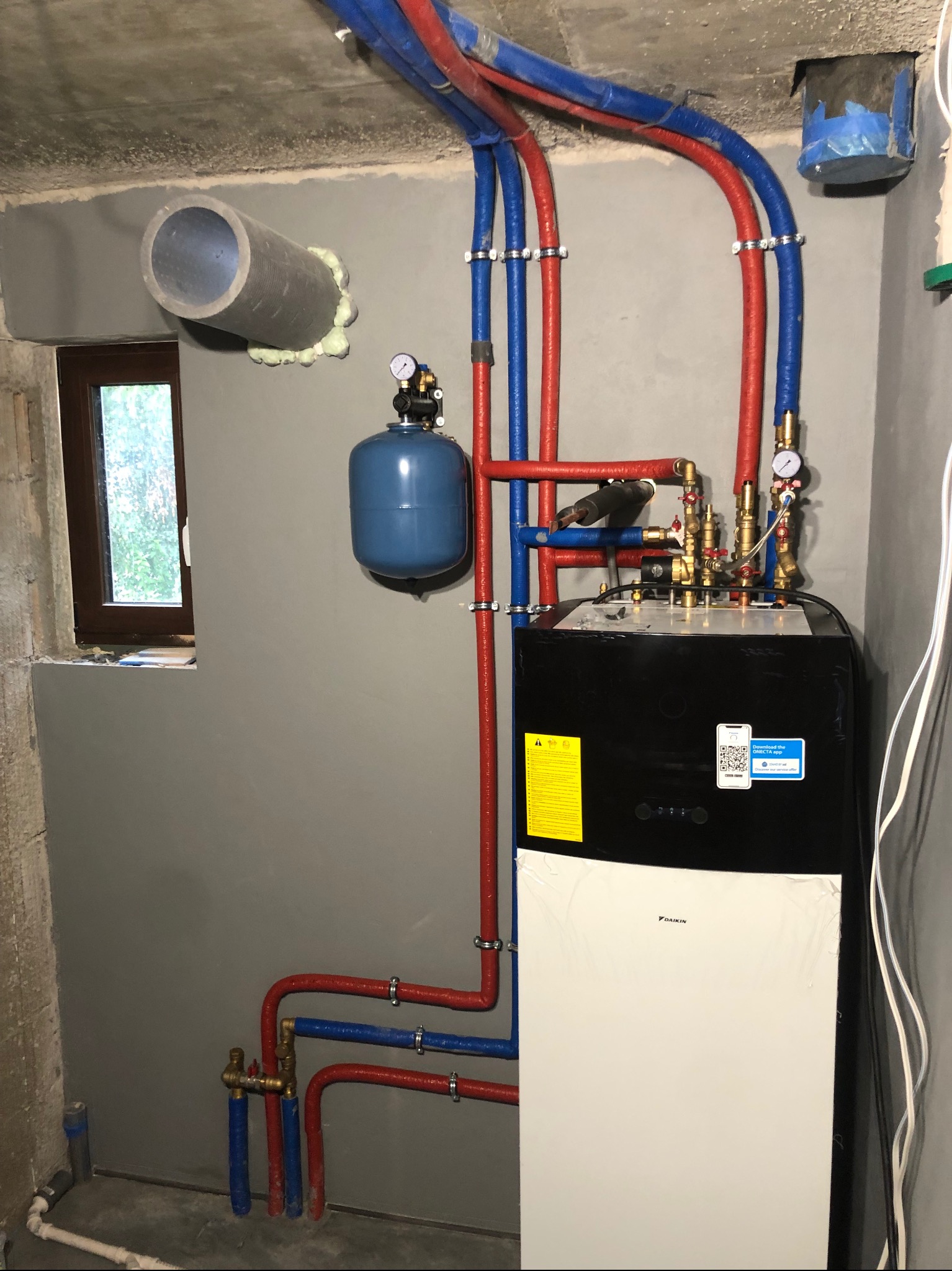 Nowoczesny system grzewczy z pompą ciepła Daikin, z widocznymi rurami w kolorze czerwonym i niebieskim, zamontowany w surowym pomieszczeniu z oknem i wystającą rurą wentylacyjną.
