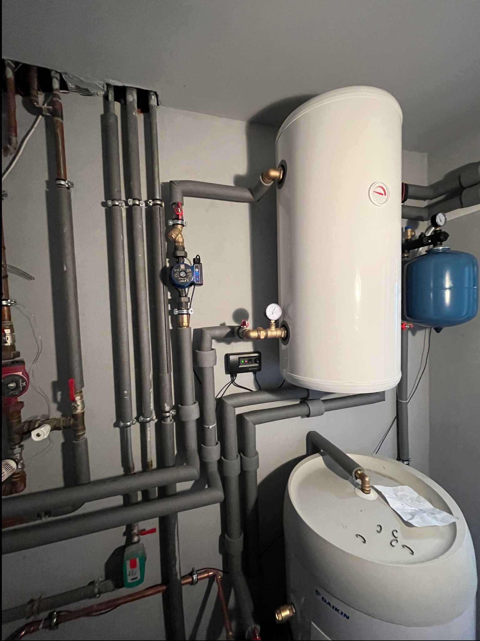 Biały zasobnik ciepłej wody użytkowej z szarą izolacją na rurach, niebieskie naczynie przeponowe, oraz instalacja hydrauliczna z zaworami i manometrami w pomieszczeniu technicznym.