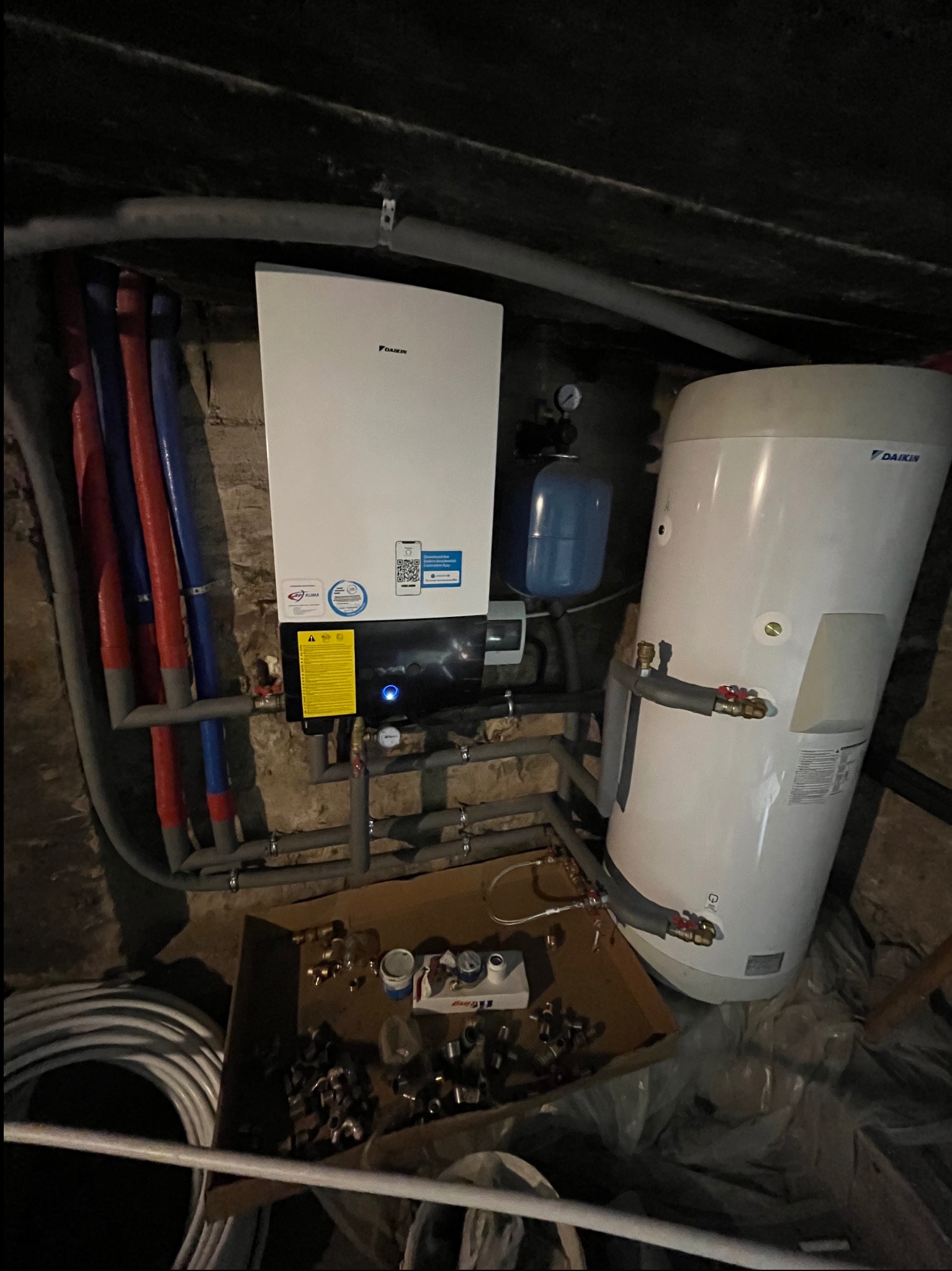 Biały piec gazowy Daikin zamontowany na ścianie, obok biały zasobnik ciepłej wody, widoczne rury instalacji grzewczej w kolorach niebieskim i czerwonym, oraz skrzynka z elementami instalacyjnymi.