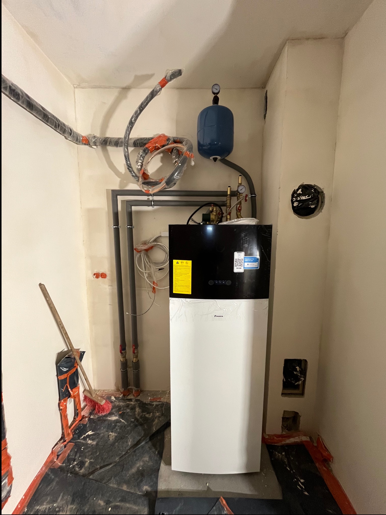 Montaż pompy ciepła Daikin w surowym pomieszczeniu w Poznaniu, widoczne rury instalacyjne zabezpieczone folią, przygotowane do podłączenia, podłoga zabezpieczona folią malarską, ściany w stanie...