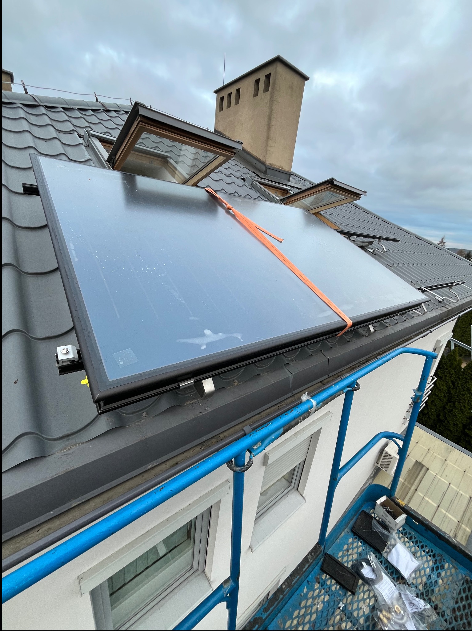 Montaż panelu słonecznego na dachu z dachówką ceramiczną, widok z podnośnika, pomarańczowy pas zabezpieczający, otwarte okna dachowe, pochmurne niebo.