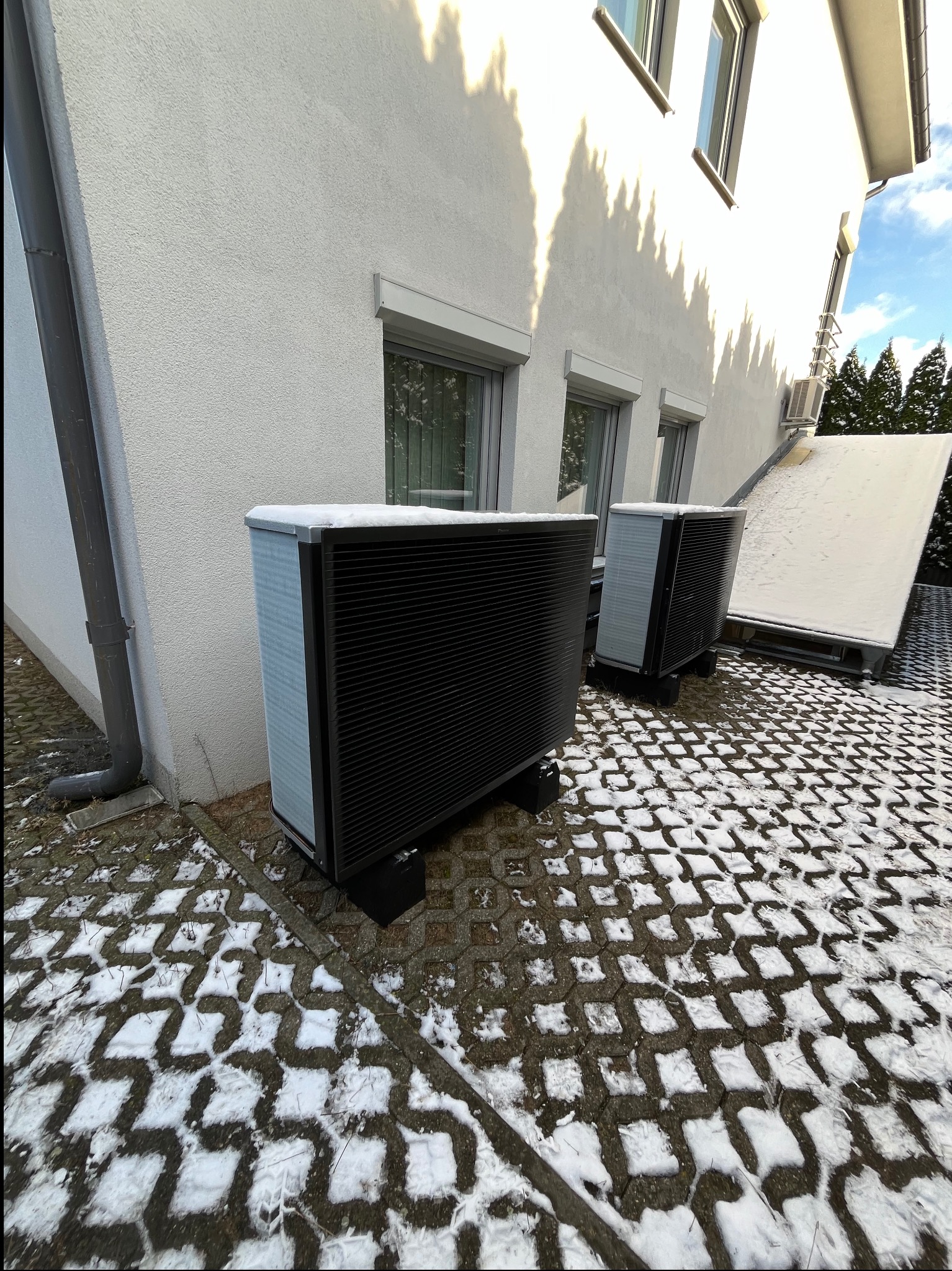 Dwie czarne pompy ciepła Daikin Altherma 3 R ECH2O, stojące na kostce brukowej pokrytej śniegiem przed jasnym budynkiem z oknami i rynną spustową.
