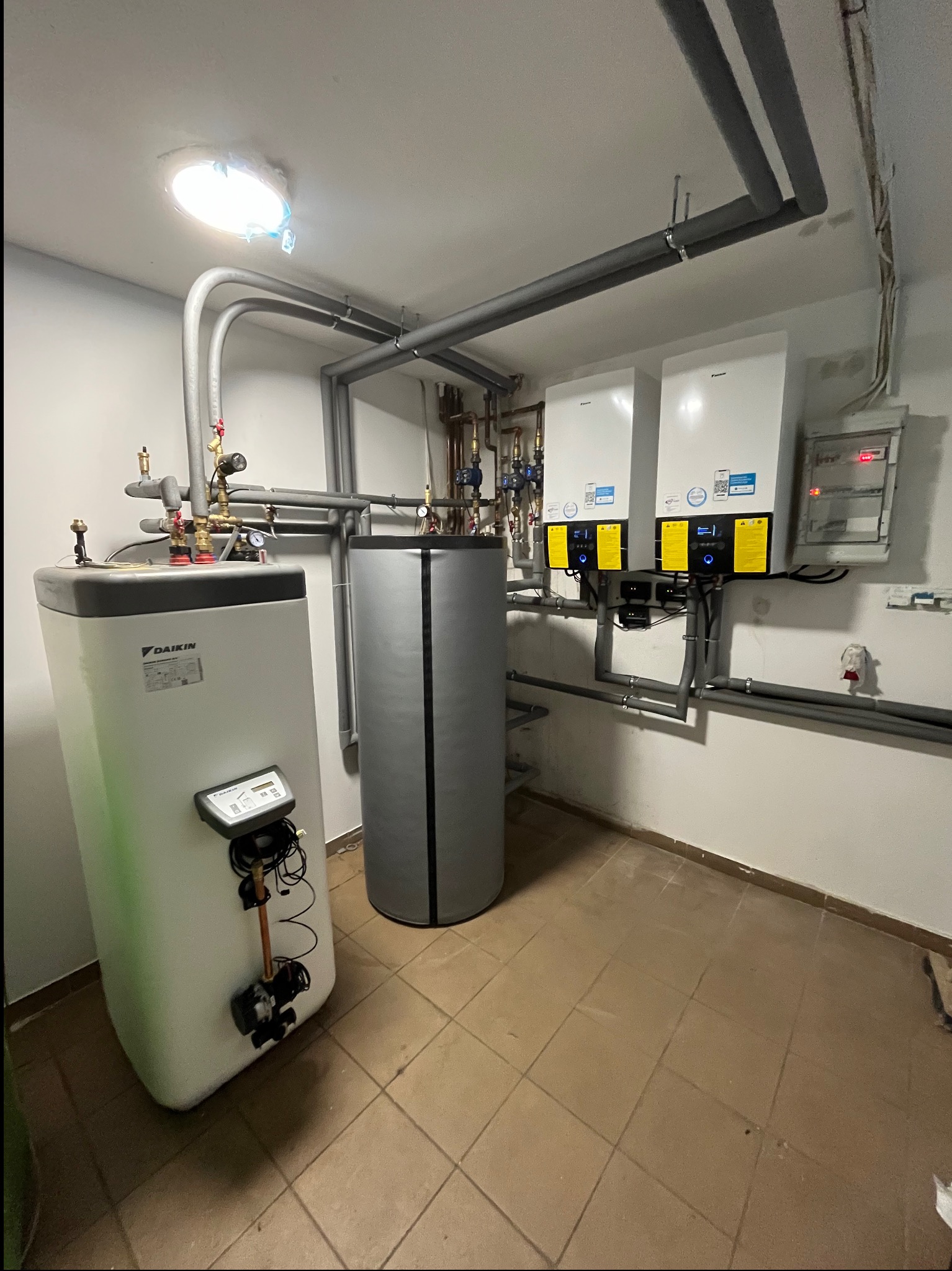 Wnętrze kotłowni z dwoma białymi jednostkami grzewczymi Daikin zamontowanymi na ścianie, obok pionowy zbiornik akumulacyjny w szarej izolacji i drugi, większy zbiornik Daikin z widocznym panelem...