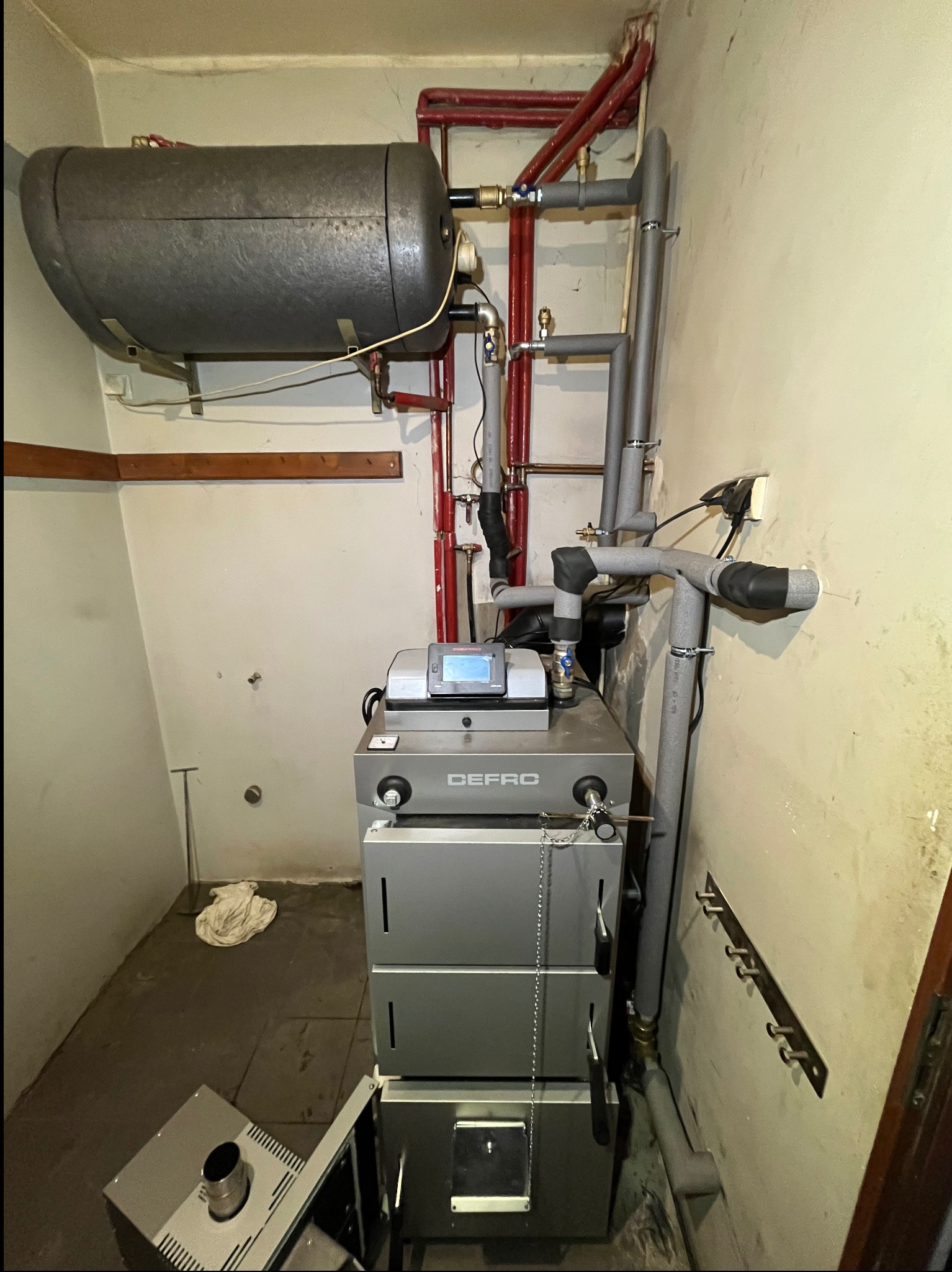 Piwnica z zainstalowanym piecem marki DEFRO, zasobnikiem wody i rurami grzewczymi, widoczne elementy instalacji CO.