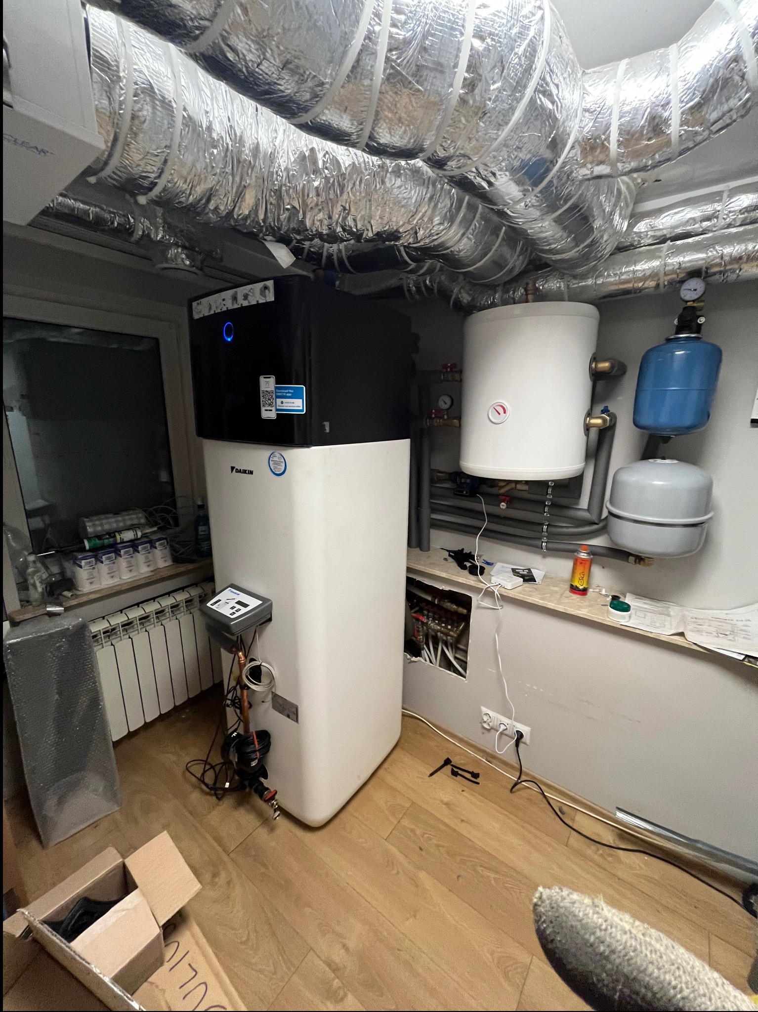 Wnętrze kotłowni z zainstalowaną pompą ciepła Daikin, zbiornikiem buforowym, naczyniami wzbiorczymi i izolowanymi rurami wentylacyjnymi pod sufitem. Widoczne także elementy instalacji hydraulicznej...