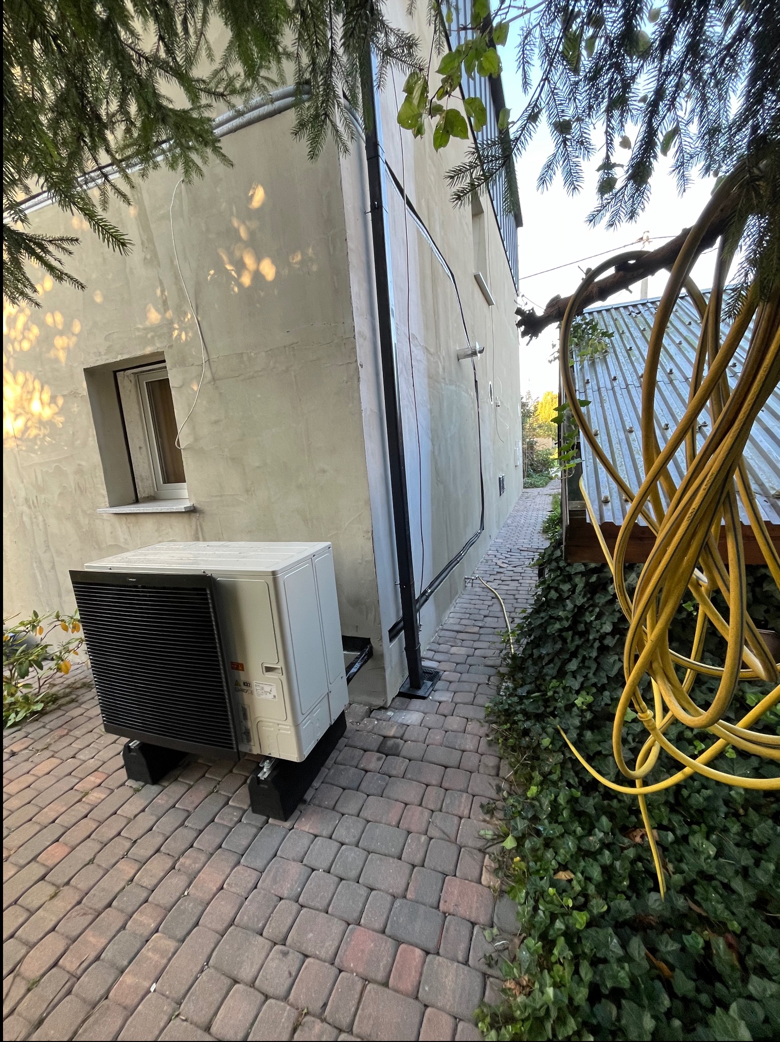 Zewnętrzna jednostka klimatyzacji lub pompy ciepła zamontowana na kostce brukowej przy budynku, widoczna z boku, z fragmentem żółtego węża ogrodowego w tle.