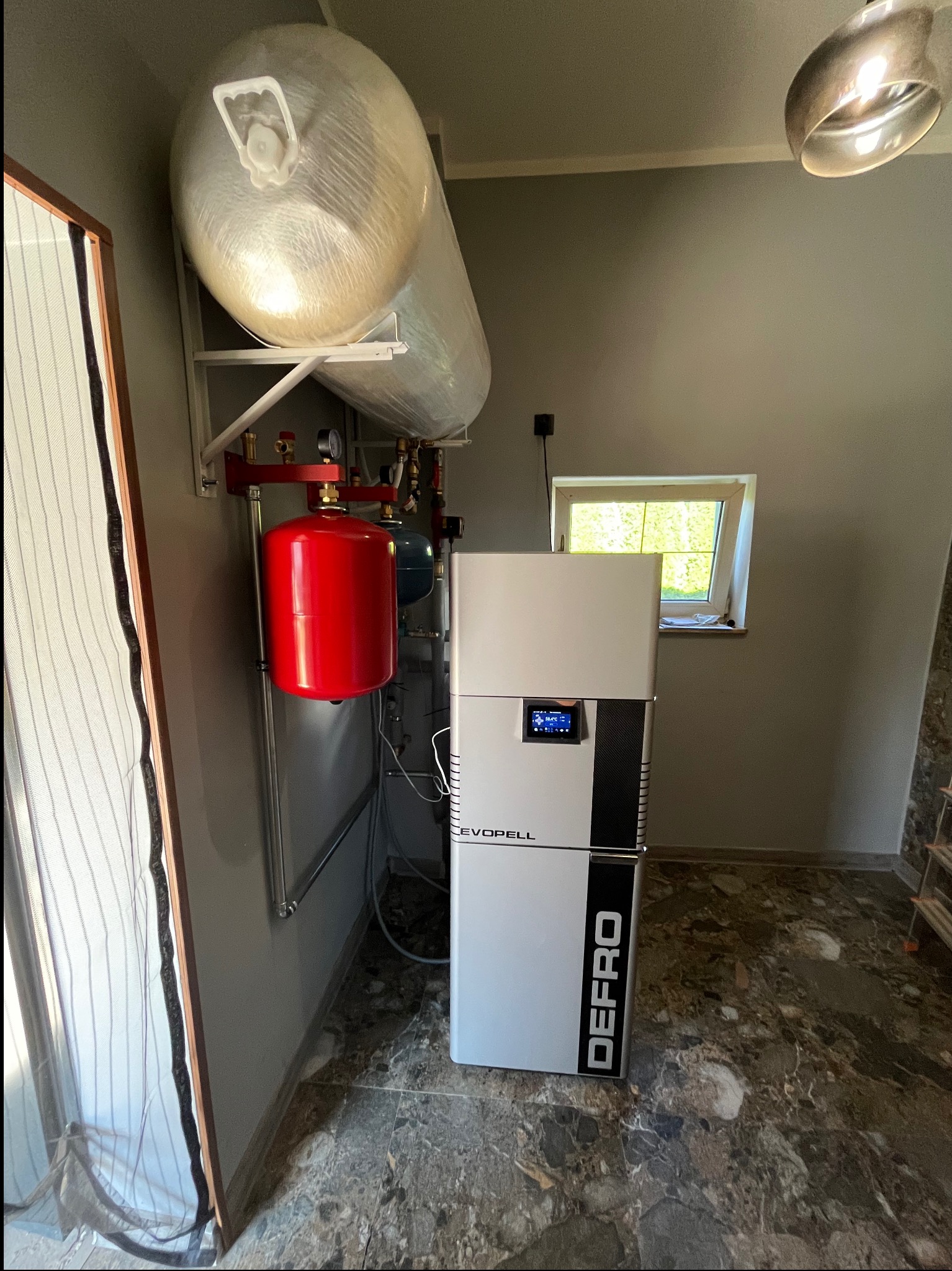 Nowoczesna kotłownia z piecem DEFRO, zbiornikiem buforowym owiniętym folią i czerwonym naczyniem przeponowym, zainstalowana w pomieszczeniu z marmurową podłogą i oknem.
