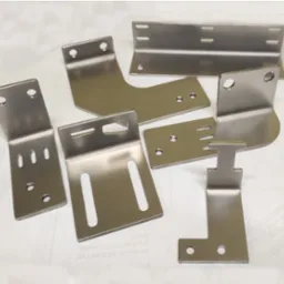 Zestaw precyzyjnie wykonanych, metalowych wspornik&oacute;w i łącznik&oacute;w o r&oacute;żnych kształtach, z otworami i szczelinami, ułożonych na białym tle z widocznym fragmentem rysunku technicznego.